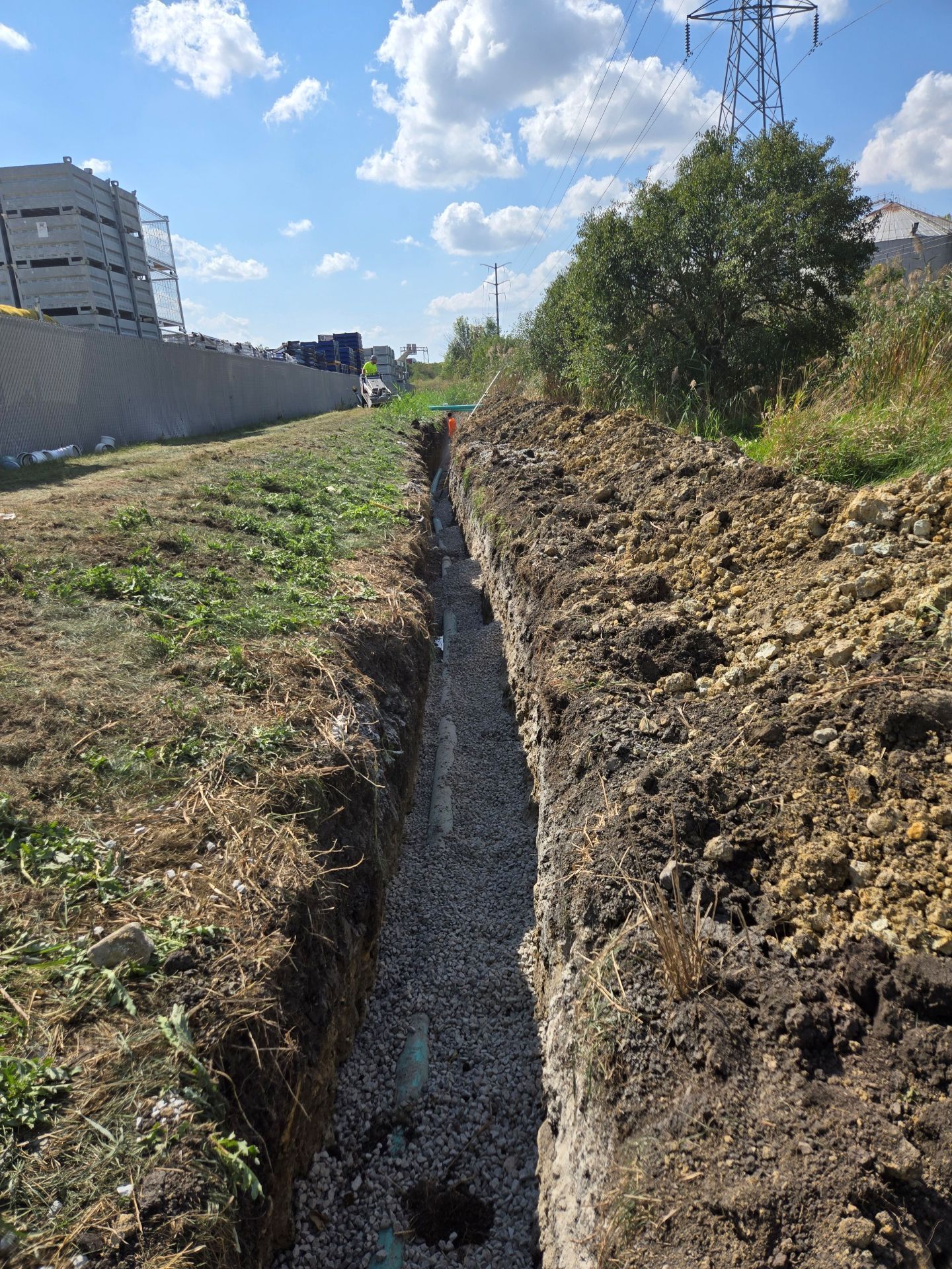 A long, narrow trench filled with gravel, alongside grassy field, under a blue sky.