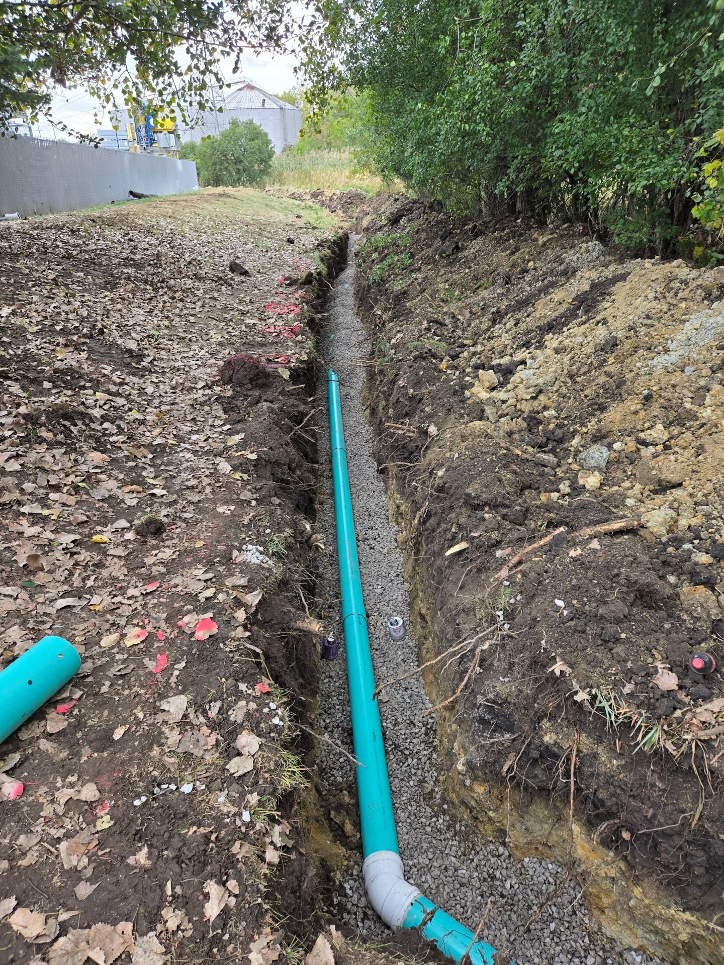 Trench with a teal pipe and gravel, likely for drainage or utilities. Dirt and vegetation surround.