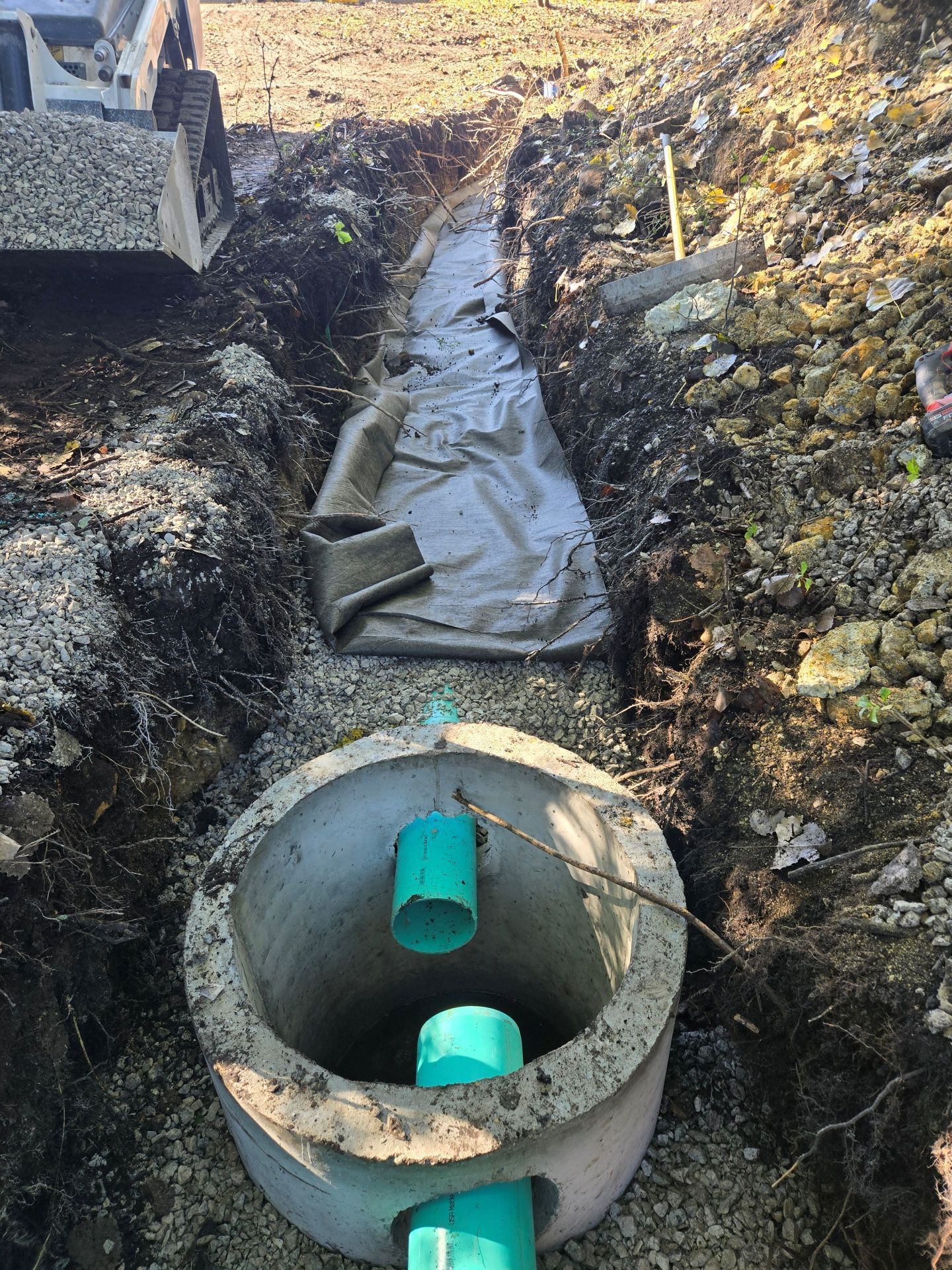 Excavated trench with concrete well and green pipes, gravel, and fabric. Earthmoving equipment in the background.