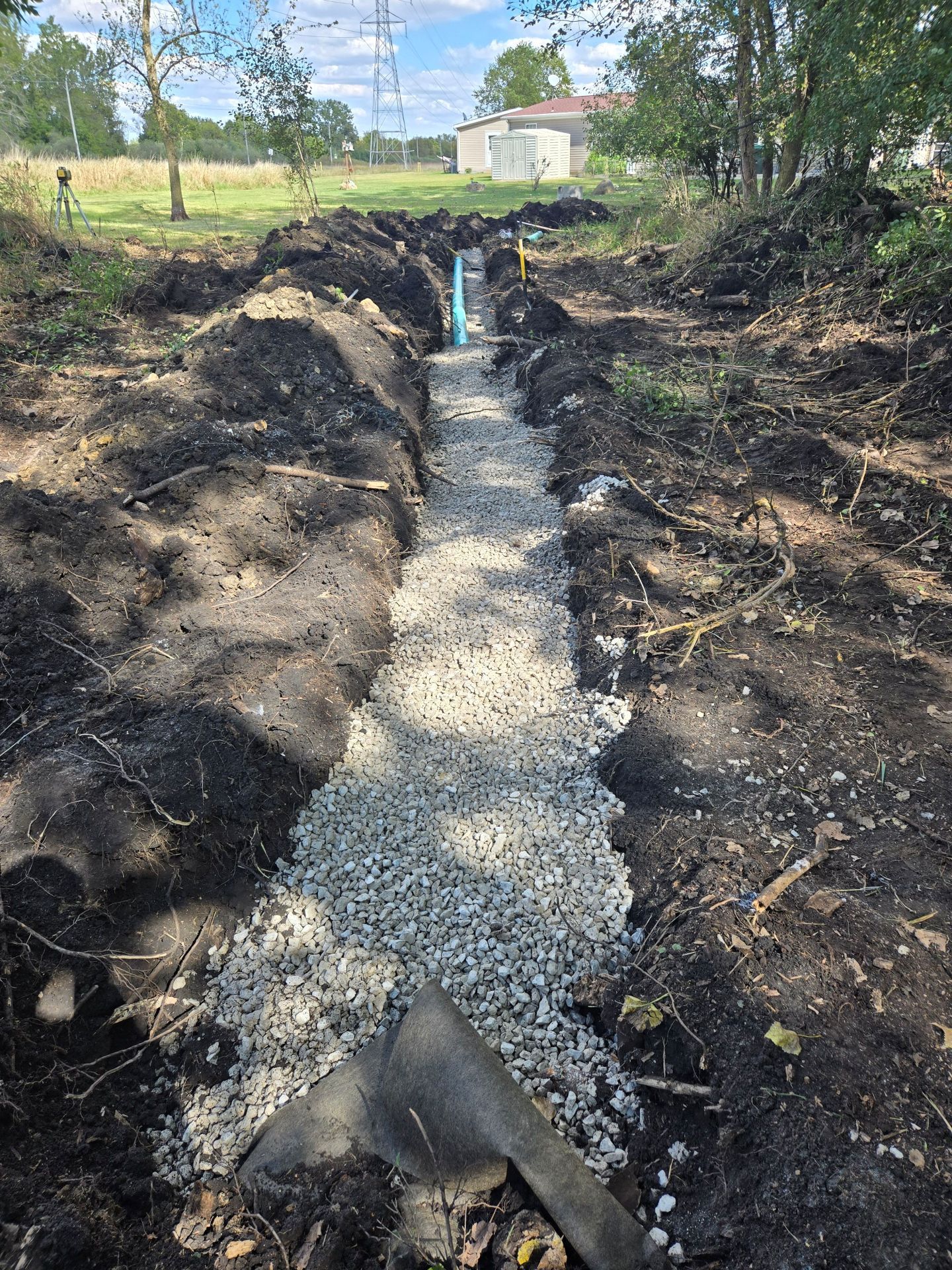 Trench filled with gravel, a blue pipe visible. Earth piled on either side. Outdoors, sunny day.