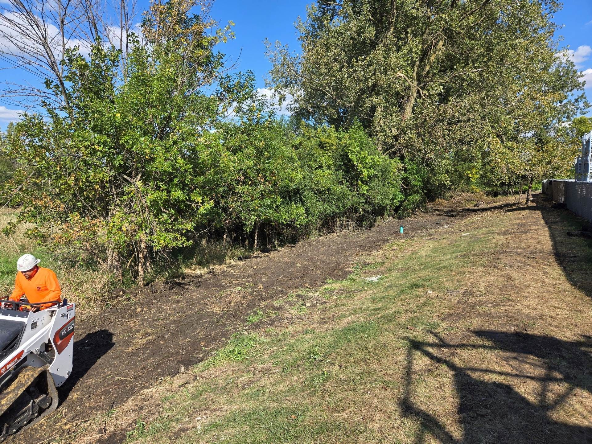 A worker operating a small excavator along a slope. Trees and blue sky in background.