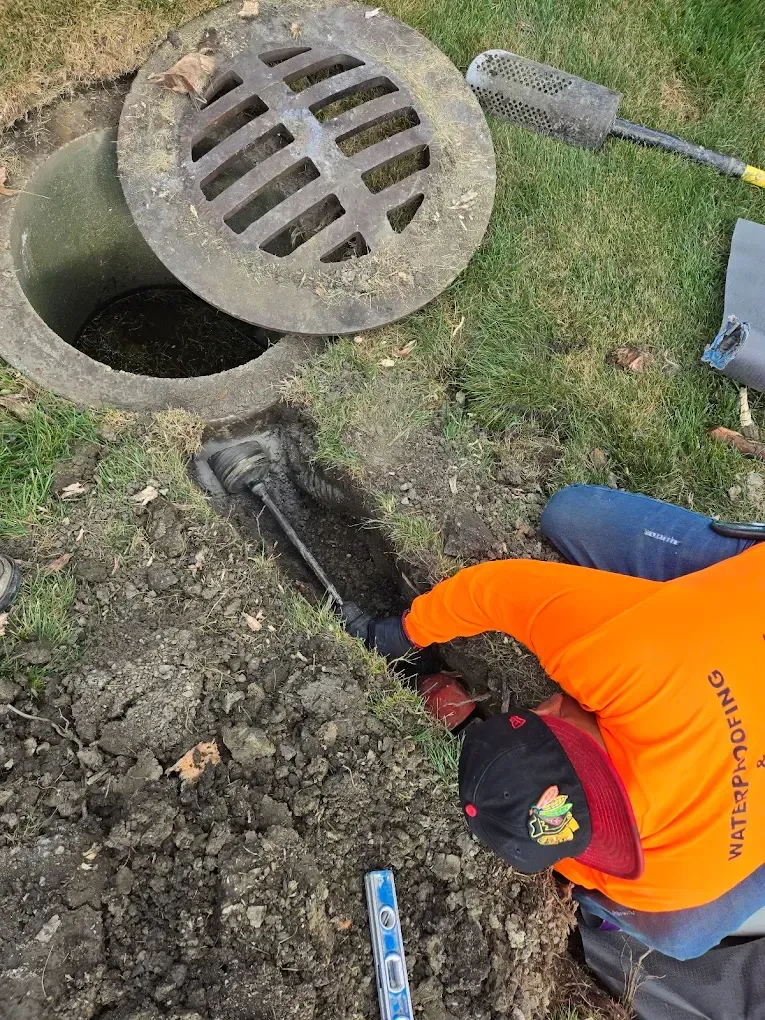 Person in orange shirt working on a drainage system, clearing a pipe with a brush.