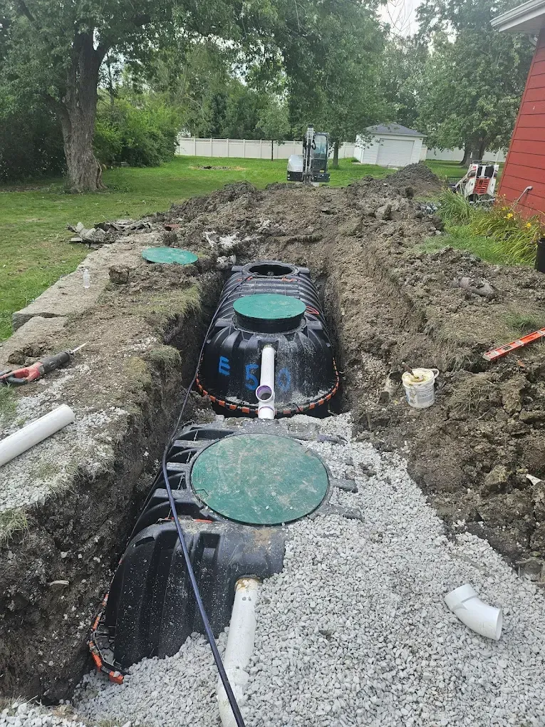 Septic system installation in progress. Two black tanks in a trench, dirt pile, and gravel.