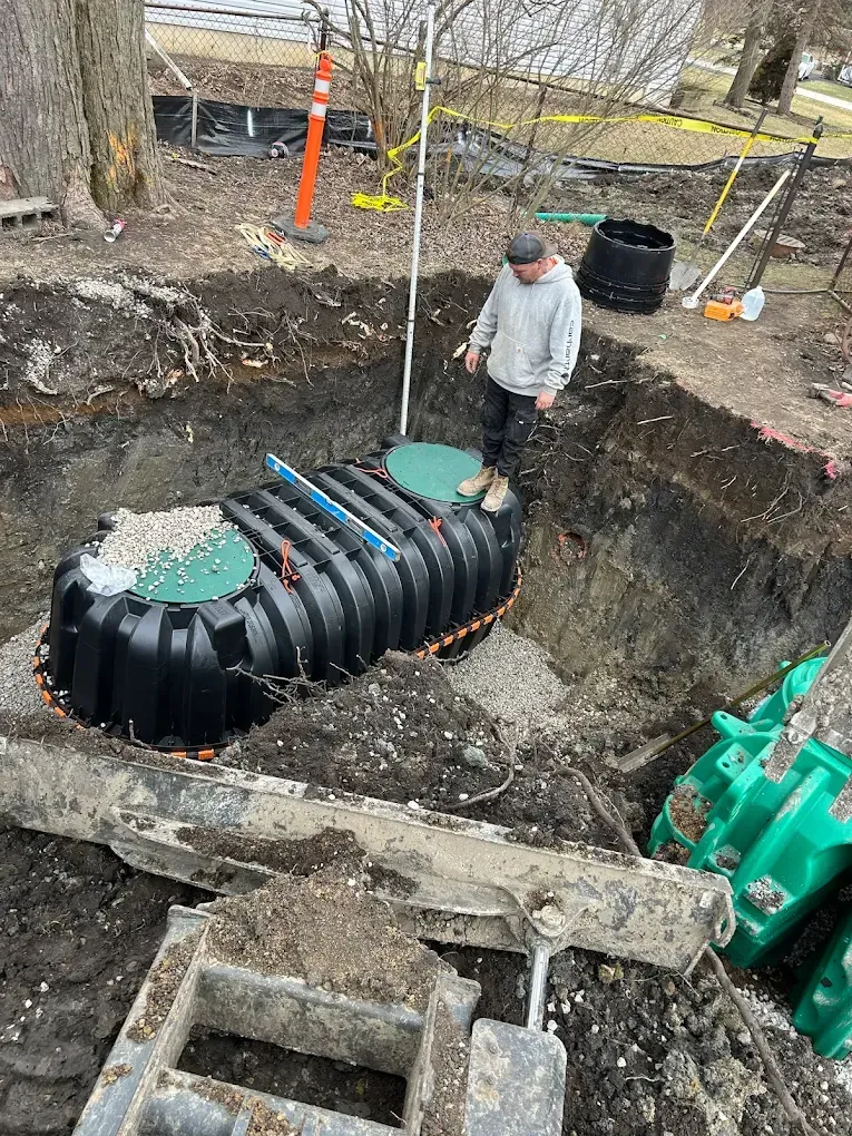 Man standing near a black septic tank in an excavated hole with construction equipment.