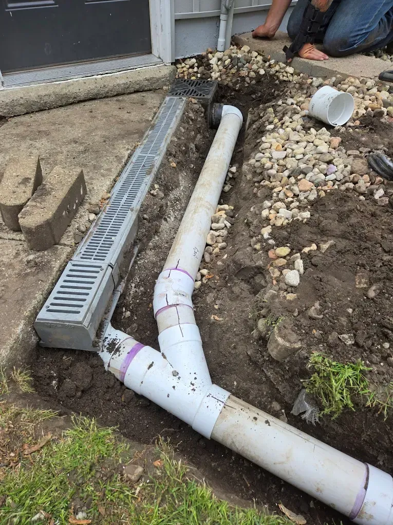 Drainage system installation: trench with gray grate, white pipes, dirt, rocks, and a person working.