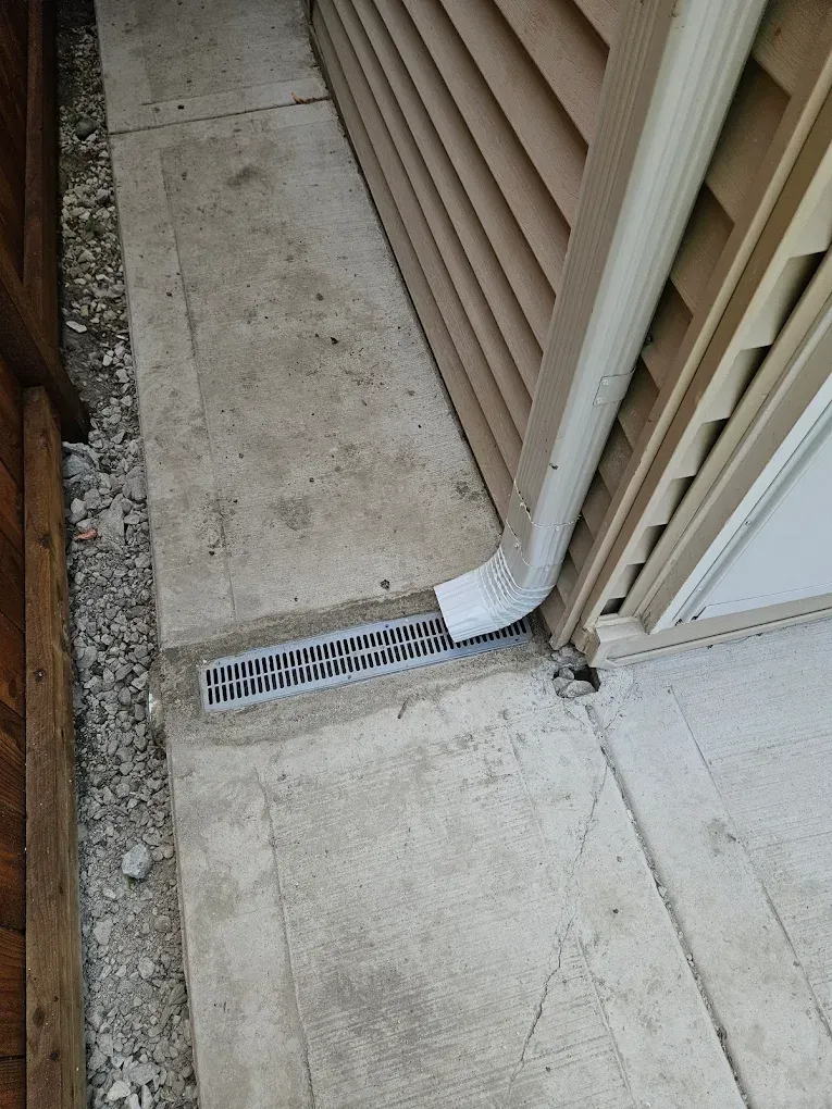 Concrete walkway with a drain under a downspout, next to a building and fence.