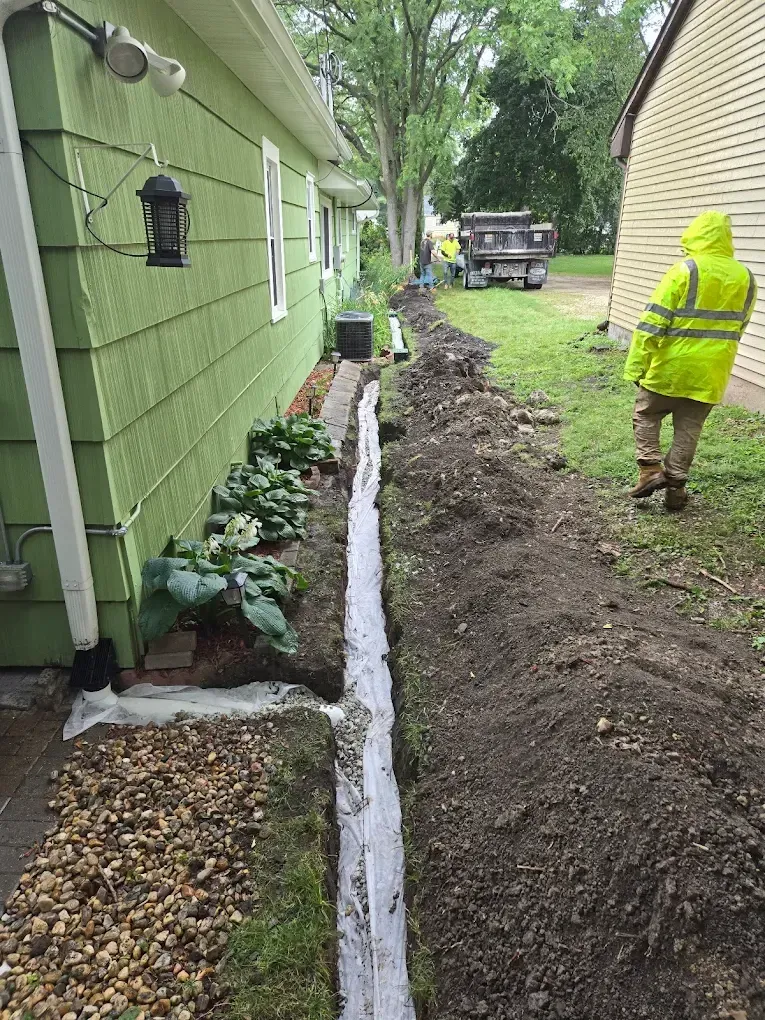 Trench dug next to a green house with a worker in yellow rain gear.