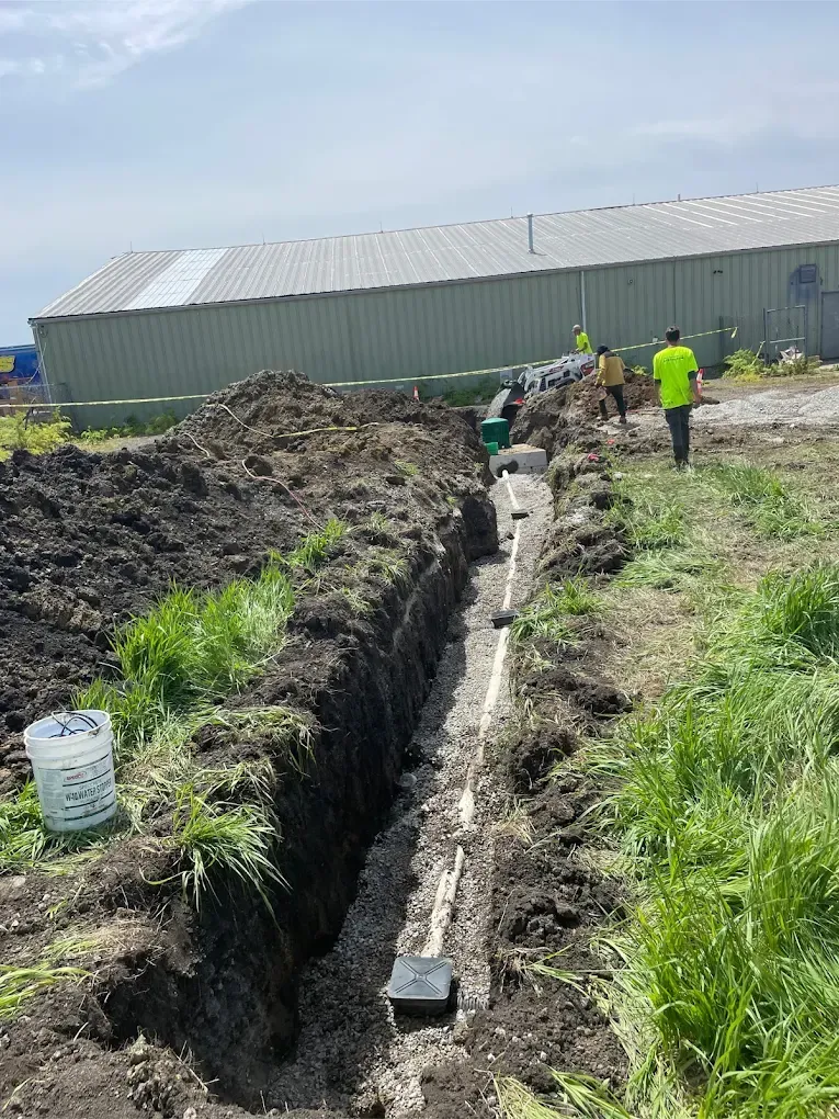 Construction workers installing underground pipes in a trench next to a building on a grassy field.