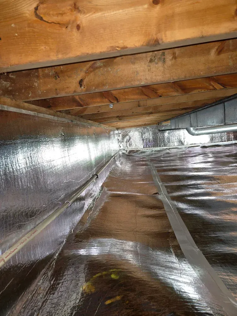 View of a crawl space with shiny insulation covering the ground and silver tape. Wooden beams above.