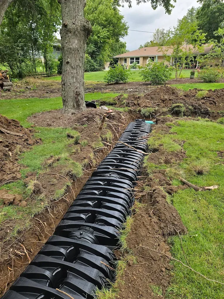 Black drainage pipe installed in a trench in a grassy yard, near a tree and a house.
