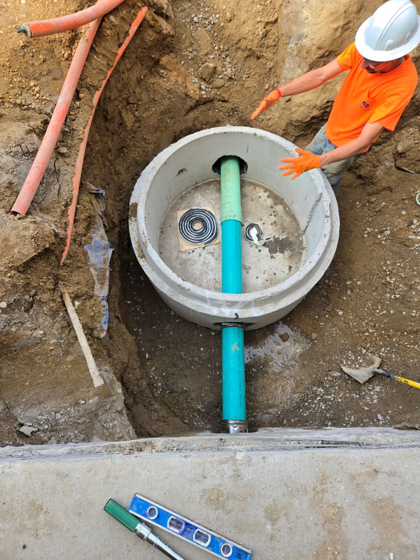 Construction worker pointing at a manhole with a pipe in a trench. Level and orange conduits are nearby.