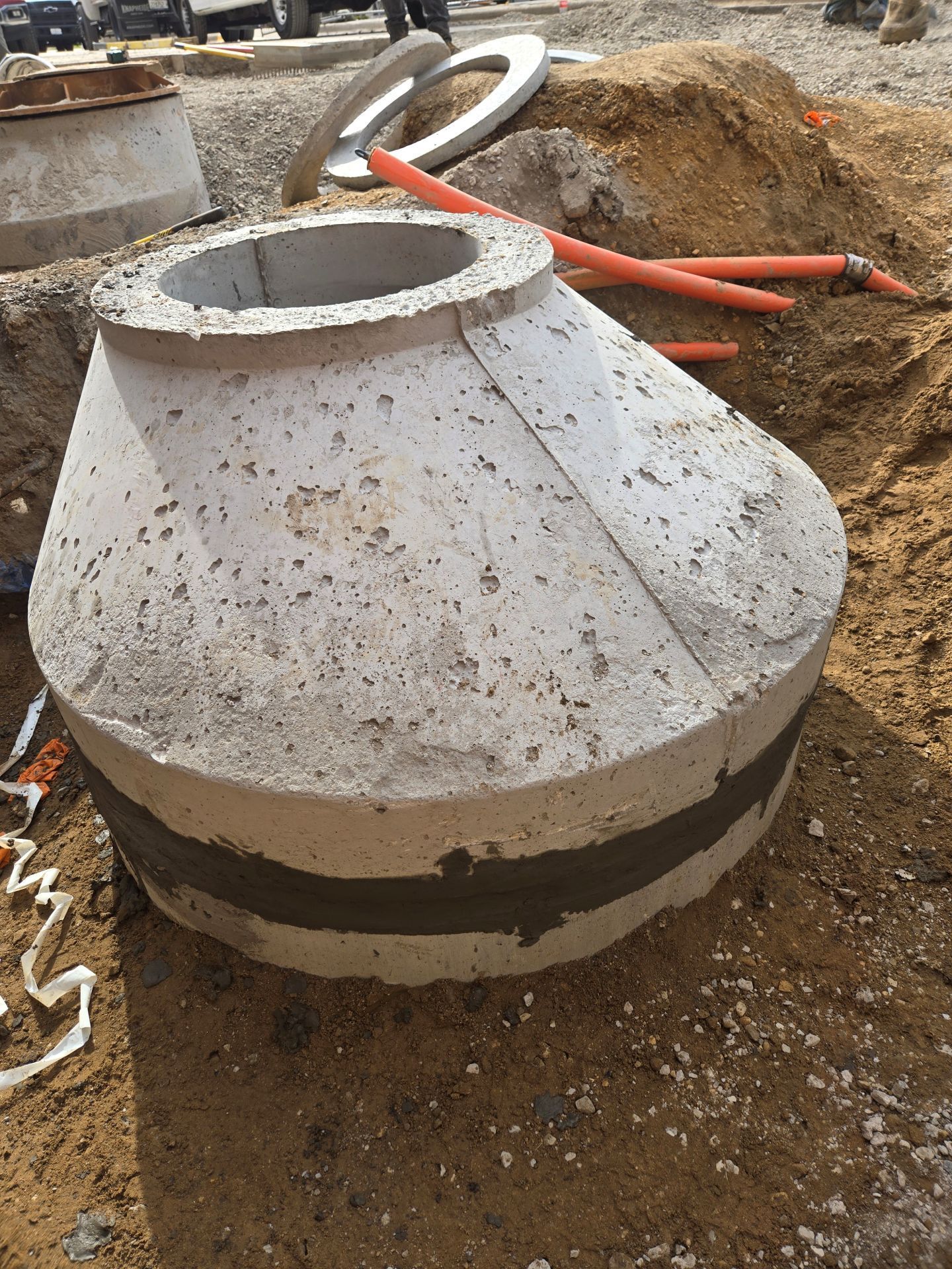 Concrete manhole cone section on a construction site, with orange pipes nearby.