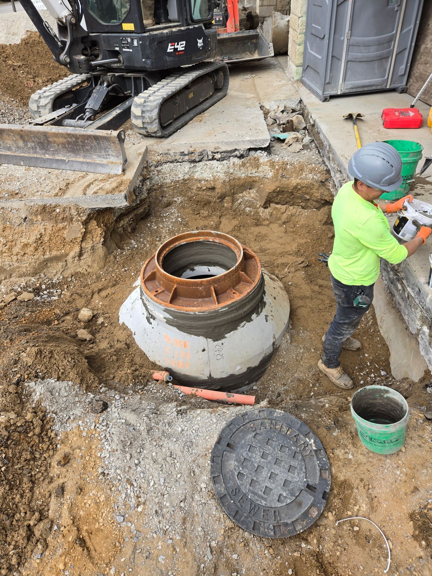 Construction worker near an open sewer access point, operating tools near a building. Excavator in the background.