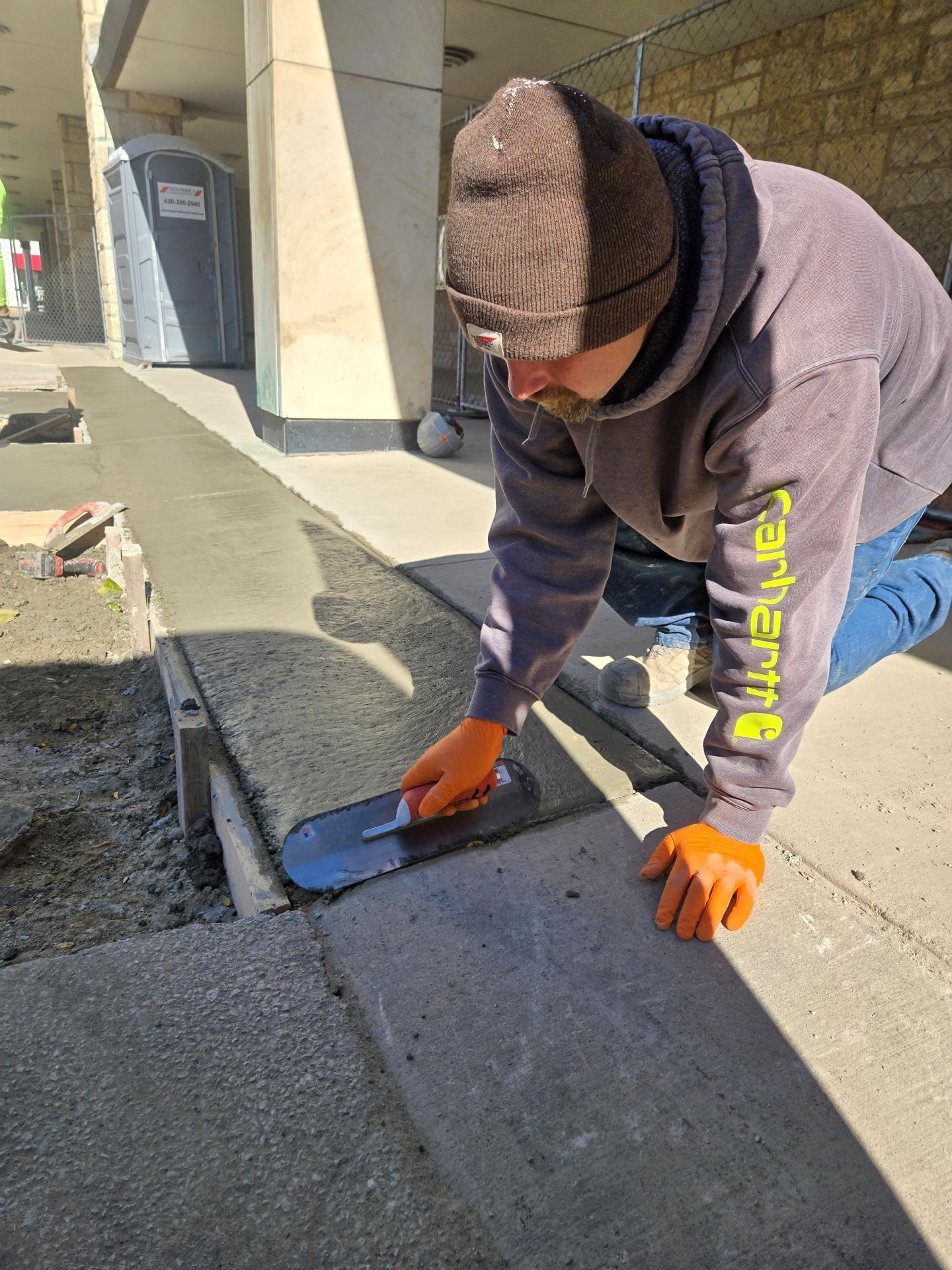 Man smoothing wet concrete sidewalk with a trowel, wearing a beanie and orange gloves.