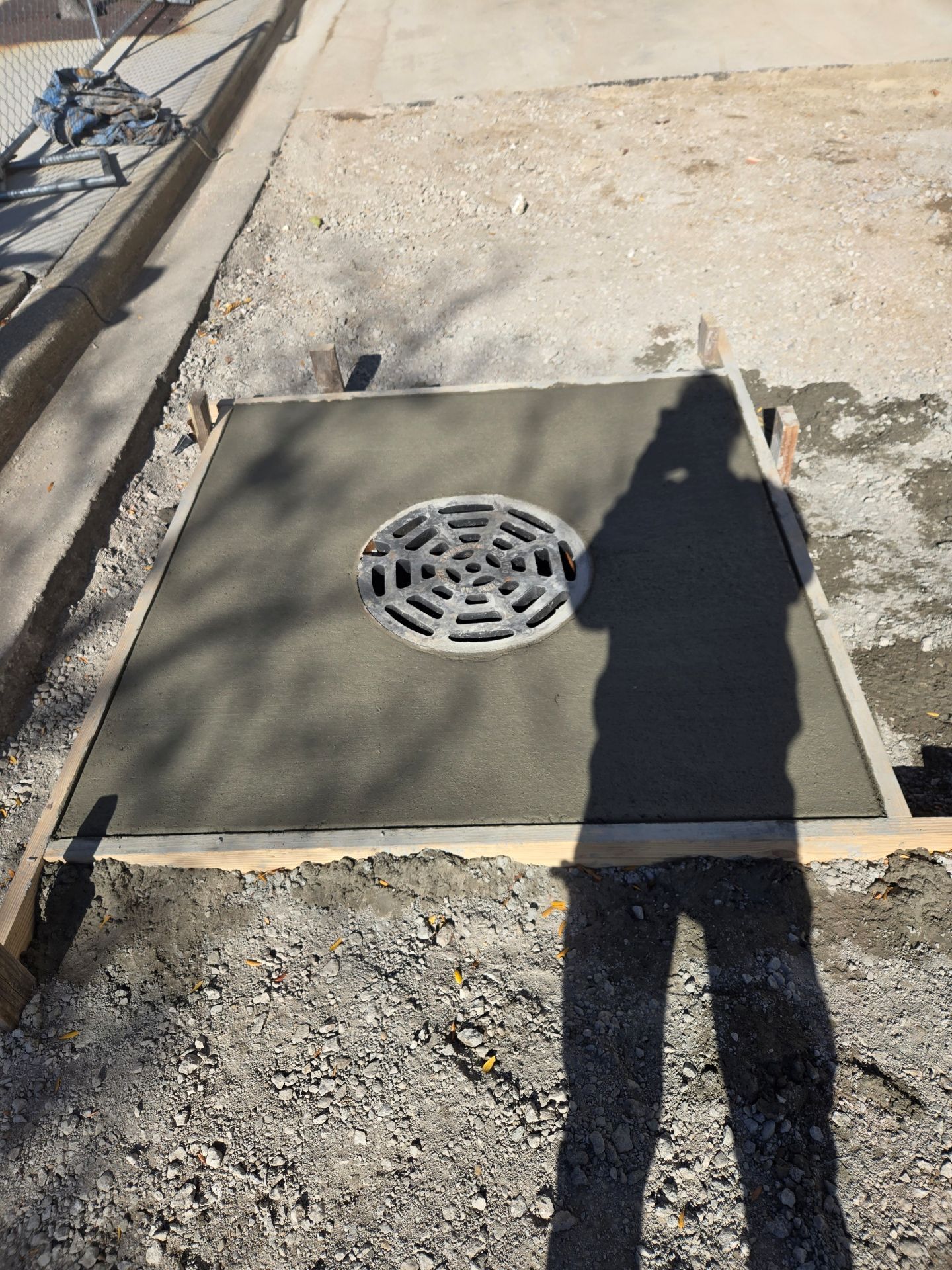 New concrete around a drain, framed by wood, surrounded by gravel. A person's shadow is cast over the concrete.