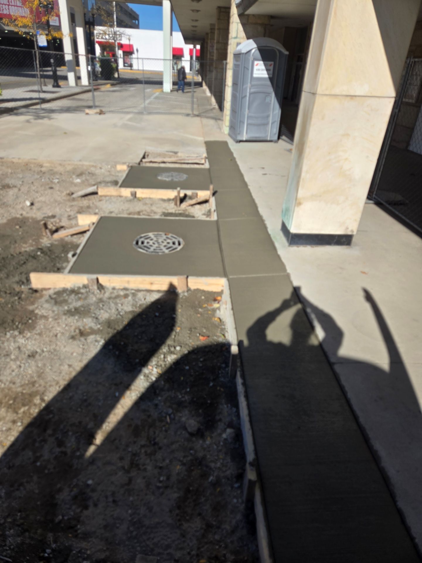 Sidewalk construction: freshly poured concrete sections with drainage grates, near a building and portable toilet.