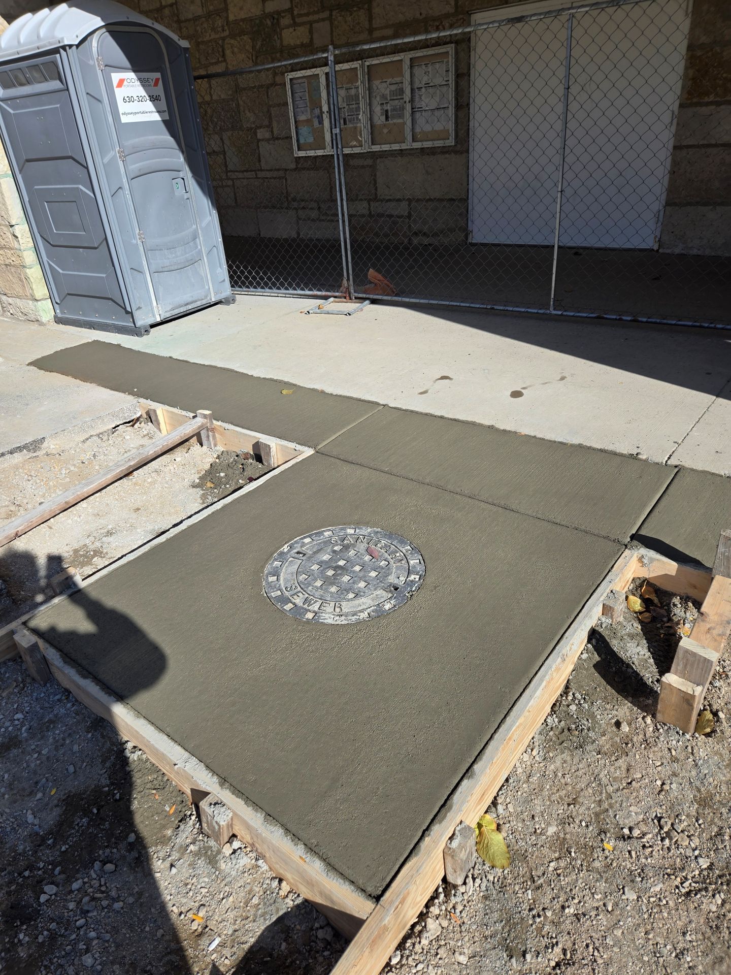 Fresh concrete sidewalk with a manhole cover, construction site, portable toilet visible.