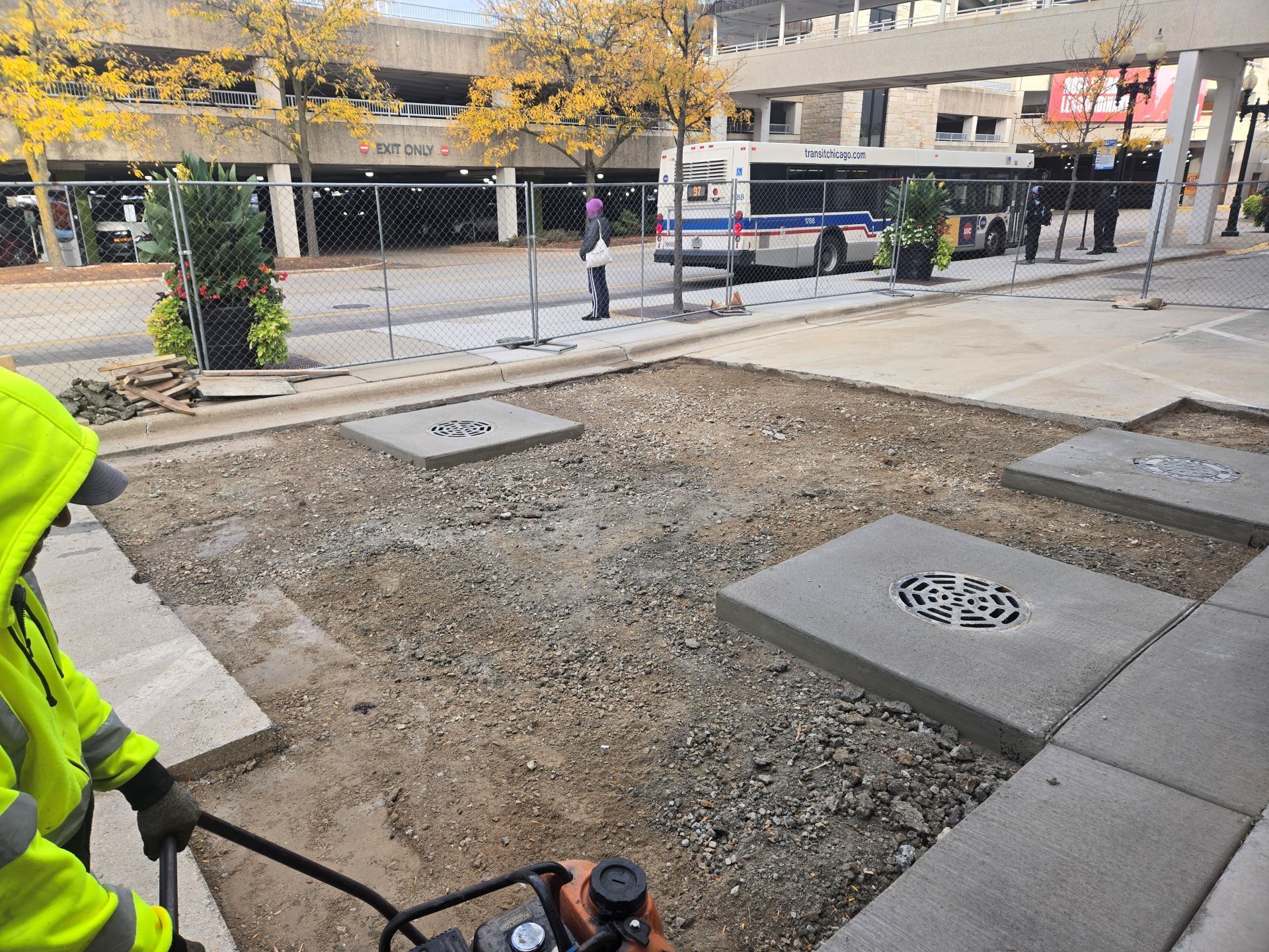 Construction site with worker compacting gravel near concrete drainage grates. Buildings and bus in background.
