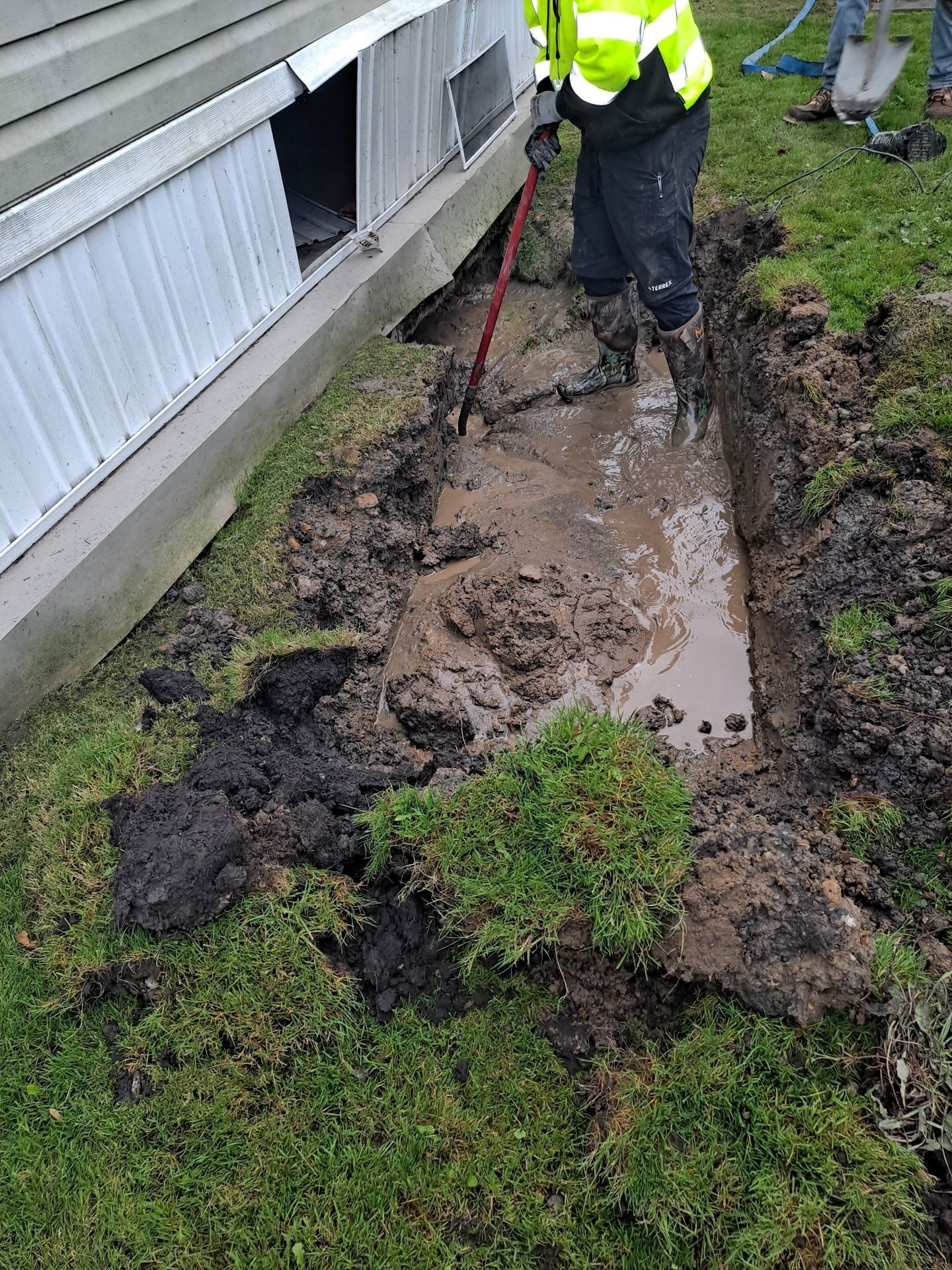 A person in a high-vis jacket digs a muddy trench along a building's foundation on a grassy lawn.