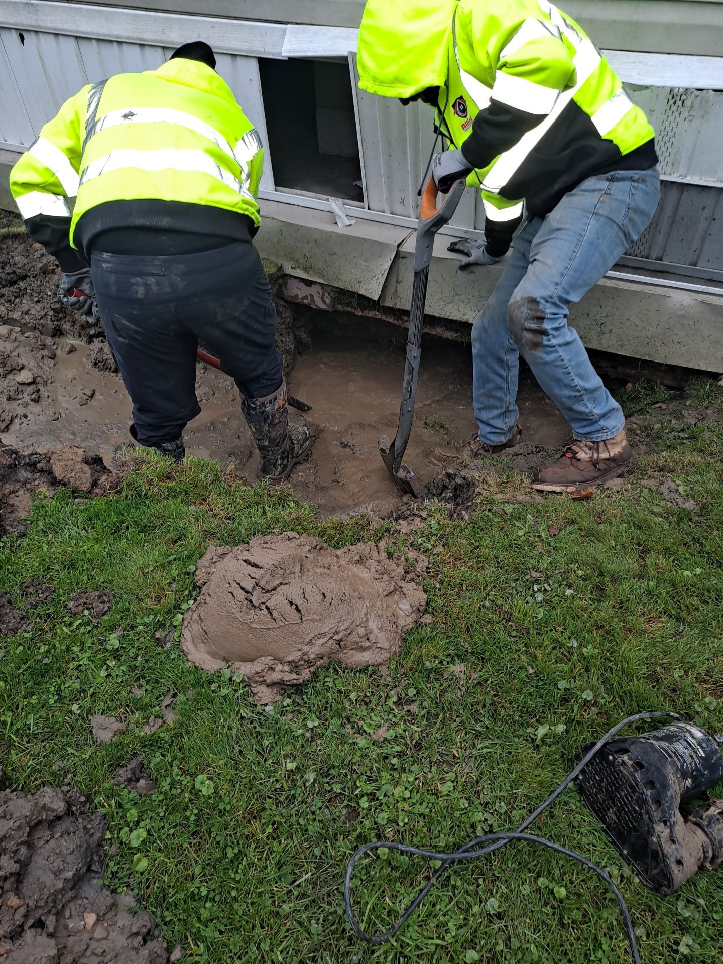 Two people in neon vests dig in muddy ground near a building.