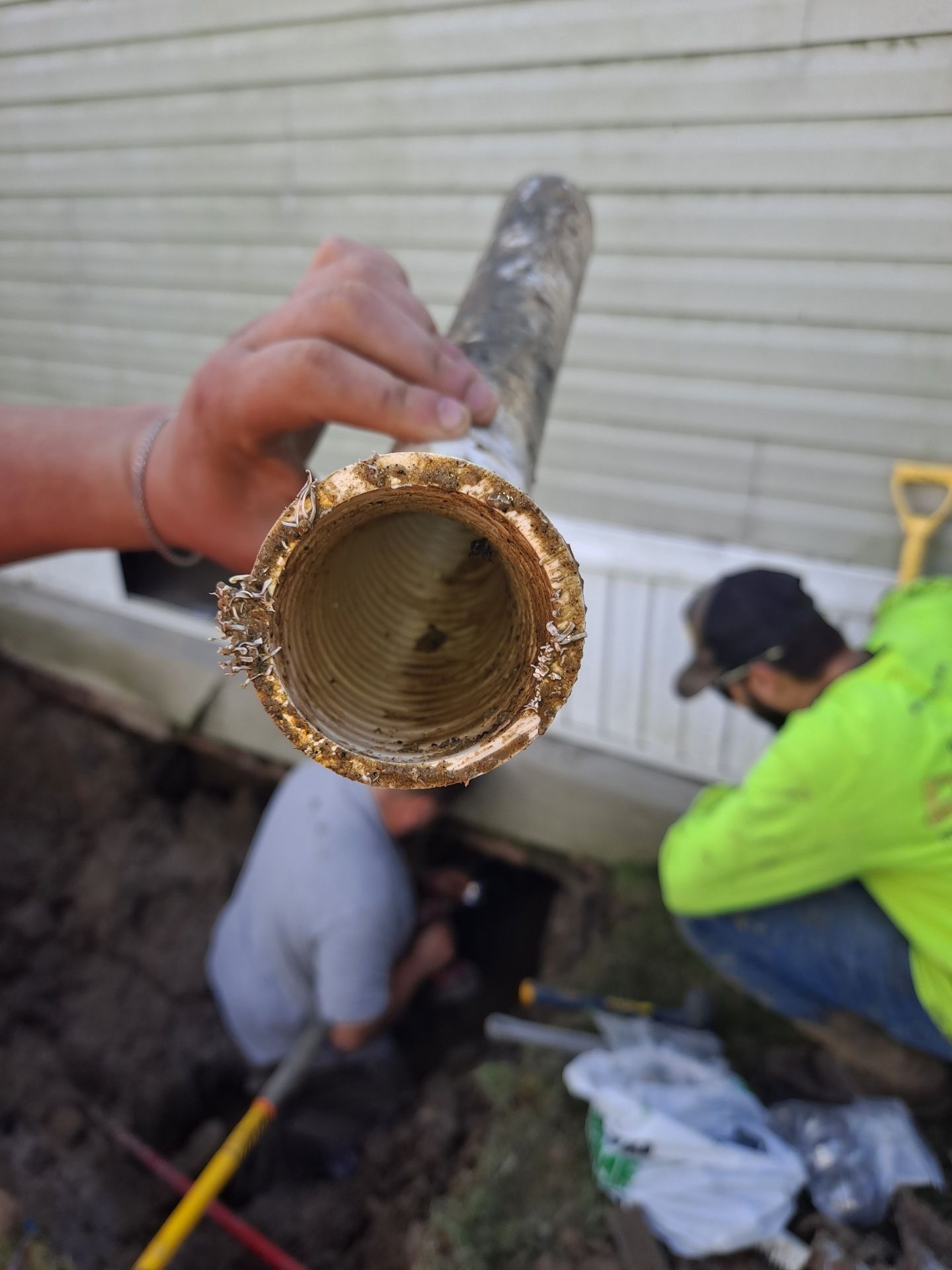 Close-up of a perforated pipe held by a person. Two workers excavate near a building.