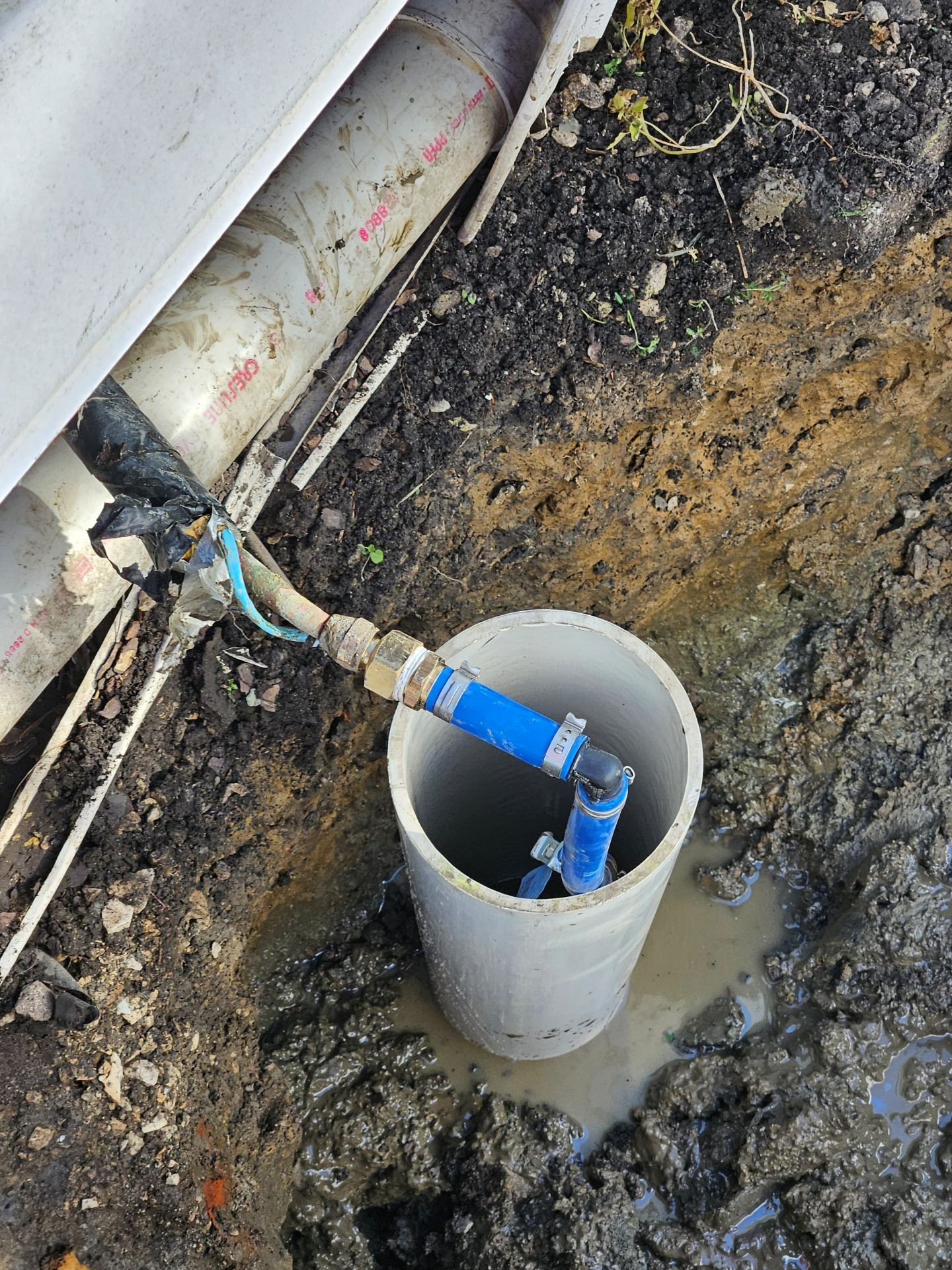 Blue plumbing fixture inside a gray pipe in a muddy hole next to other pipes and debris.