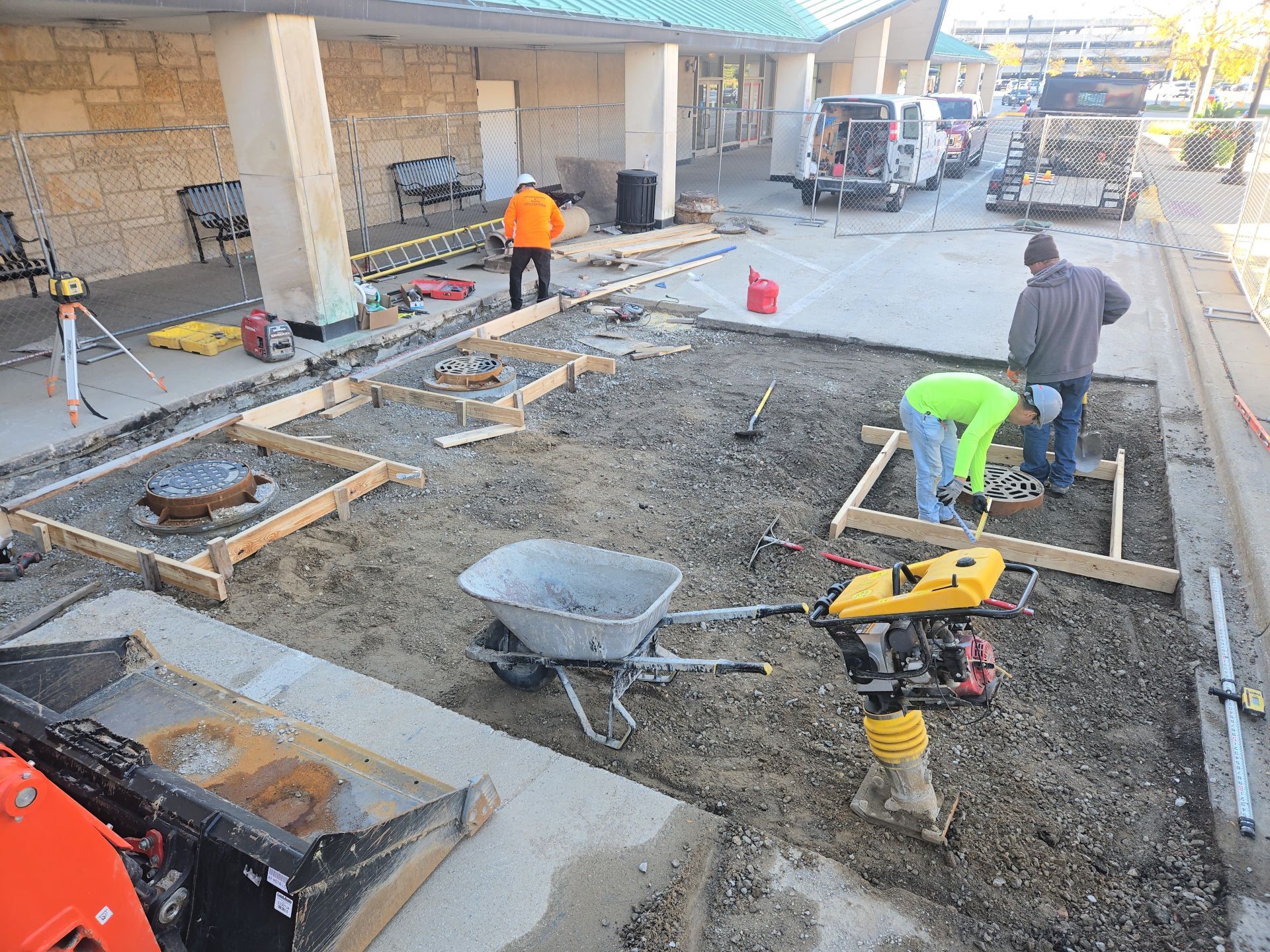 Construction workers building a sidewalk. They are using forms, a compactor, and a wheelbarrow. Outdoors.