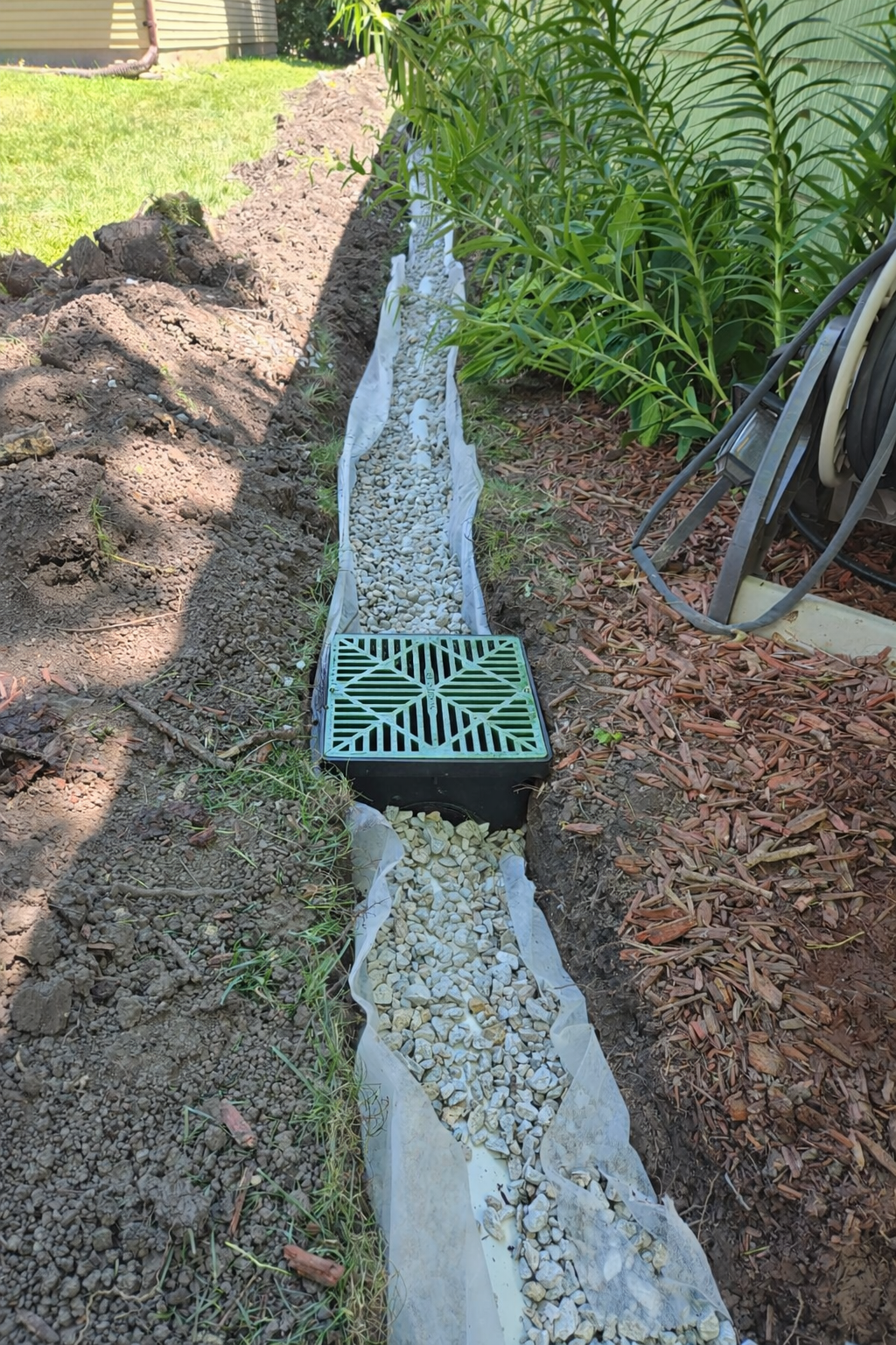 Stormwater drainage system installation with catch basin, gravel trench, and filter fabric along residential property.