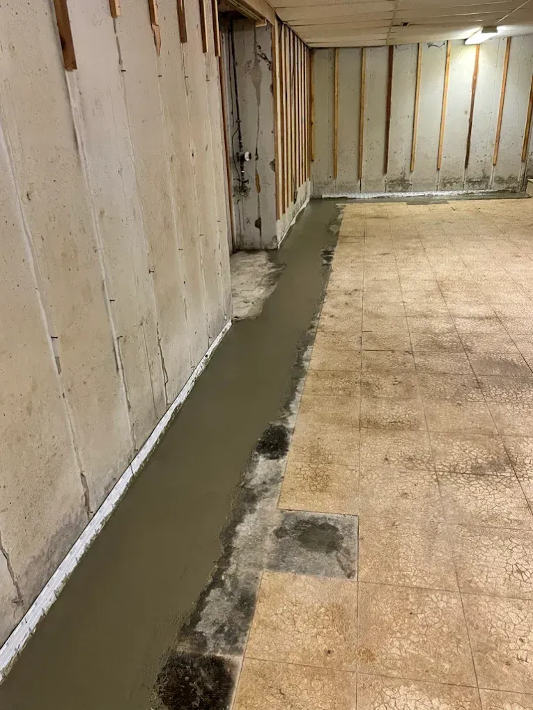 Concrete floor with a new cement strip along the wall. Basement setting, with white walls and wooden framing.