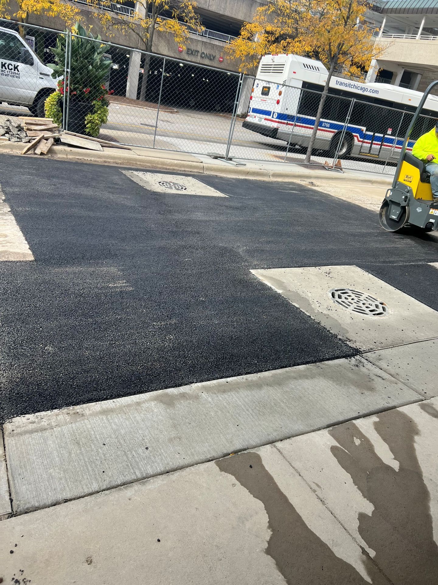 Asphalt paving in progress on a city street. A bus and construction equipment are visible.