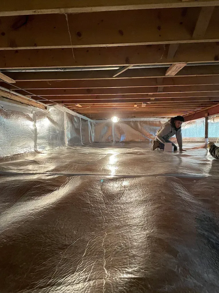 Person crawls on reflective insulation under wooden structure.