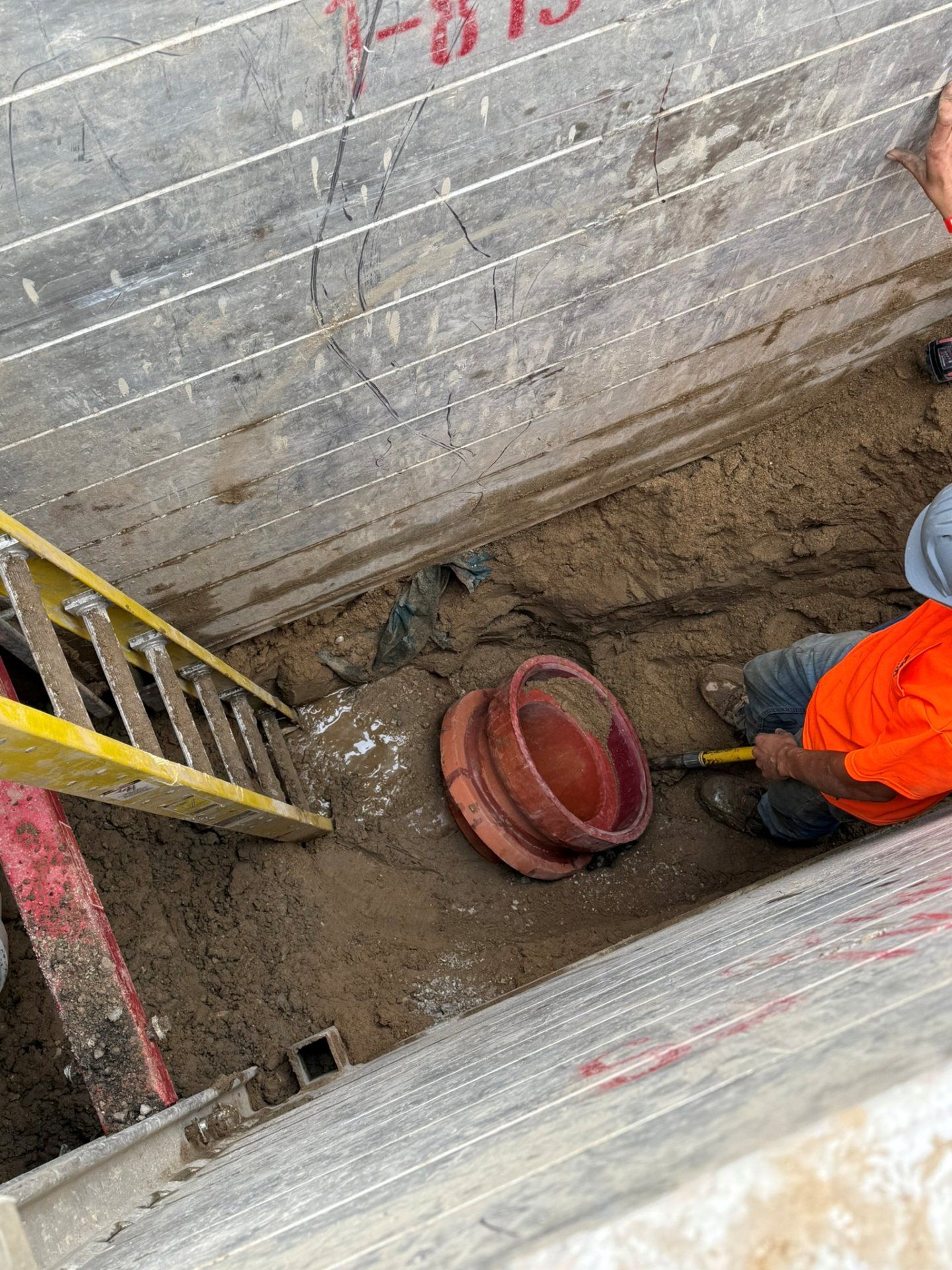 Construction worker in trench with pipe, ladder, and concrete wall.