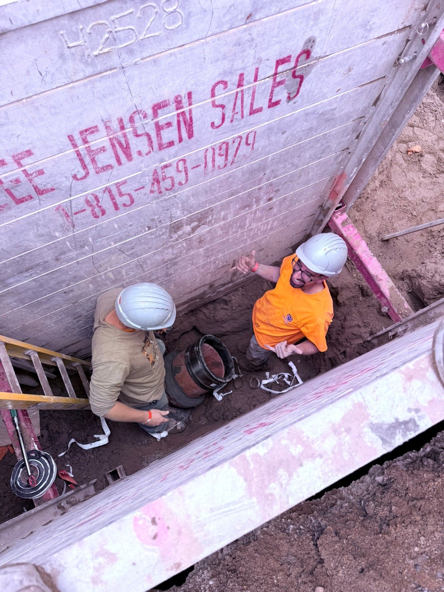Two workers in hard hats inside a trench; one looks up, other works on a pipe. Beige, orange shirts. Wooden wall.
