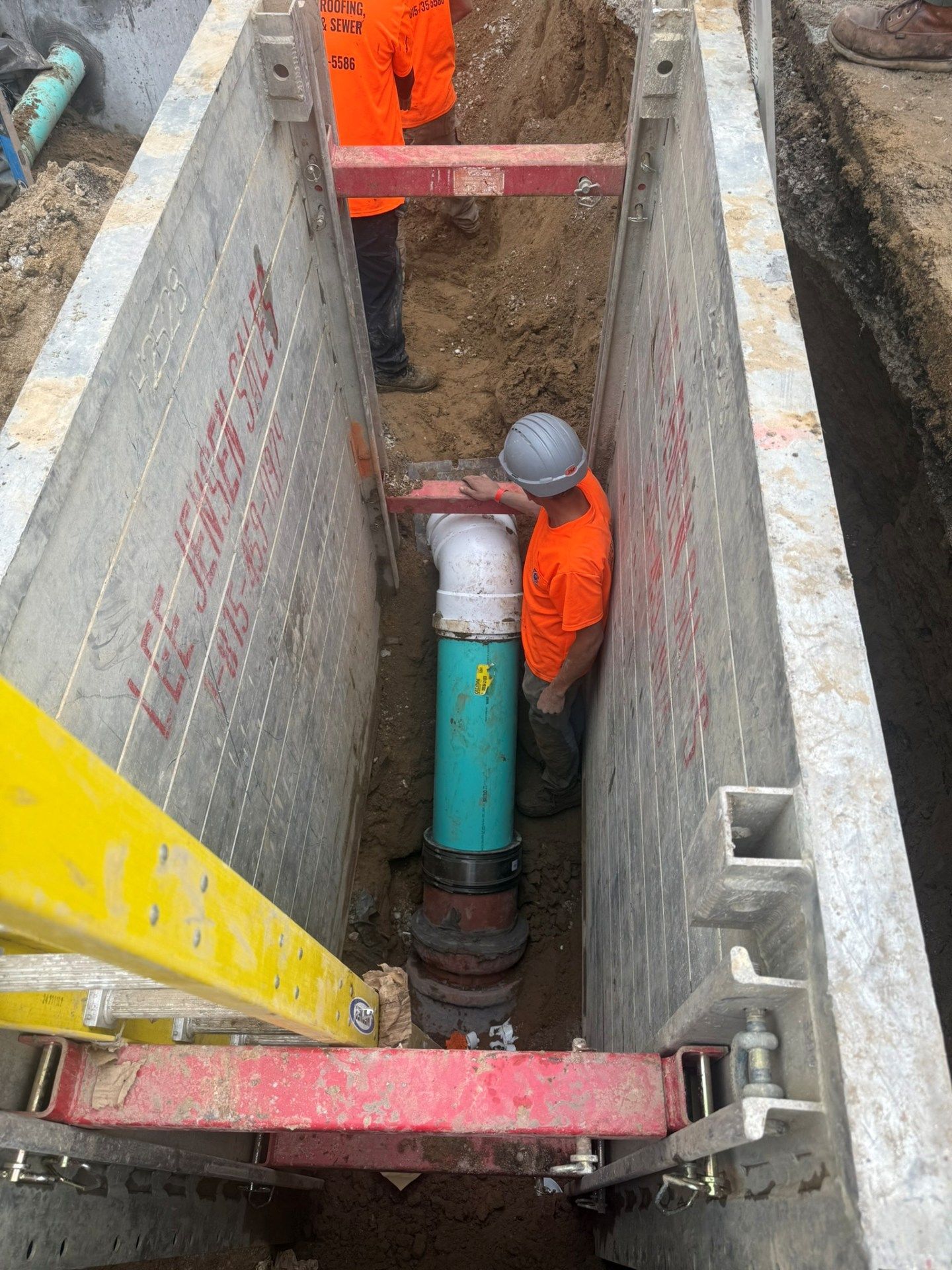 Workers in orange vests and hard hats repairing a pipe in a trench with safety supports.