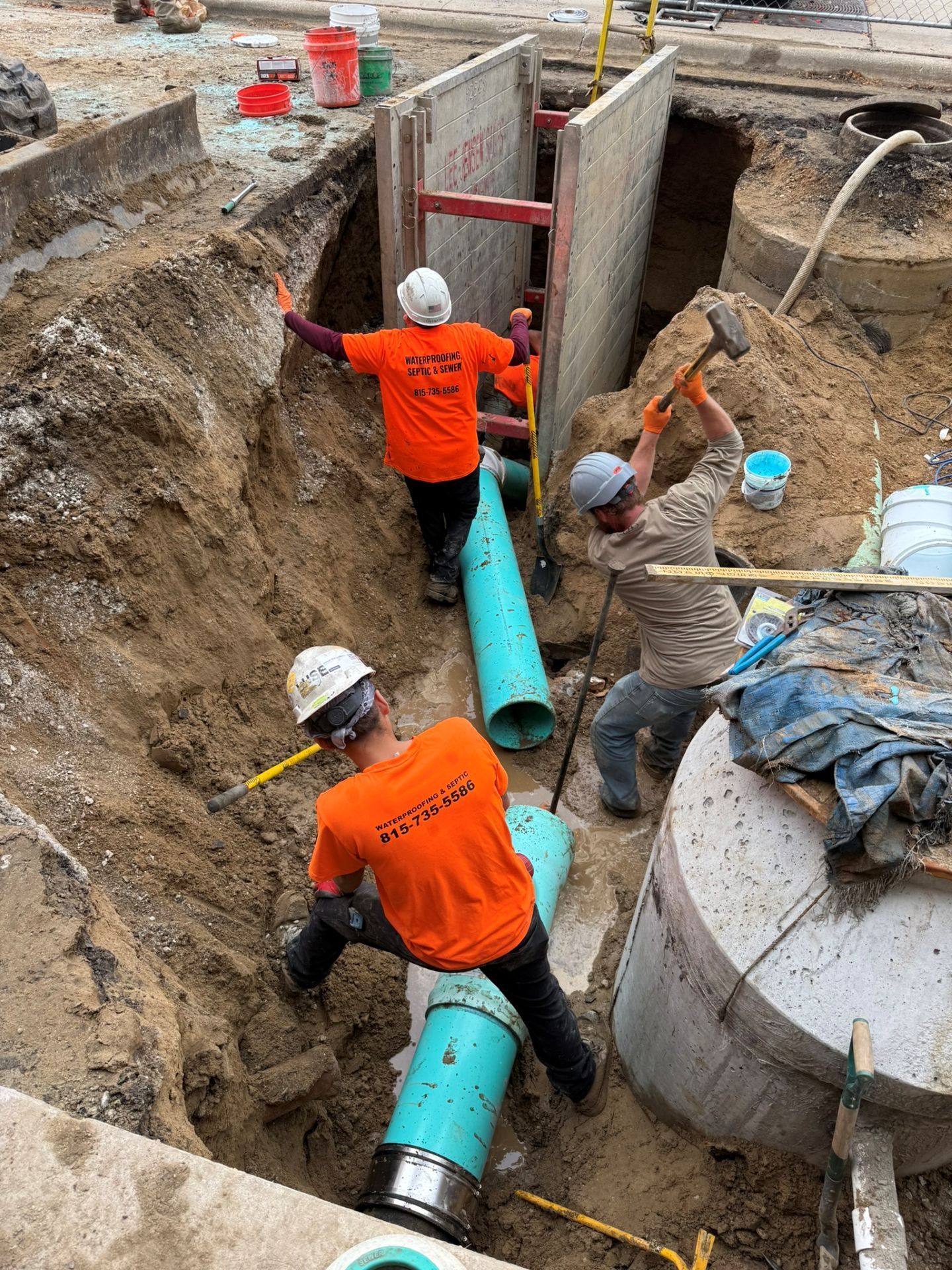 Construction workers installing large blue pipes in a trench. One swings a hammer, others work on the pipe.