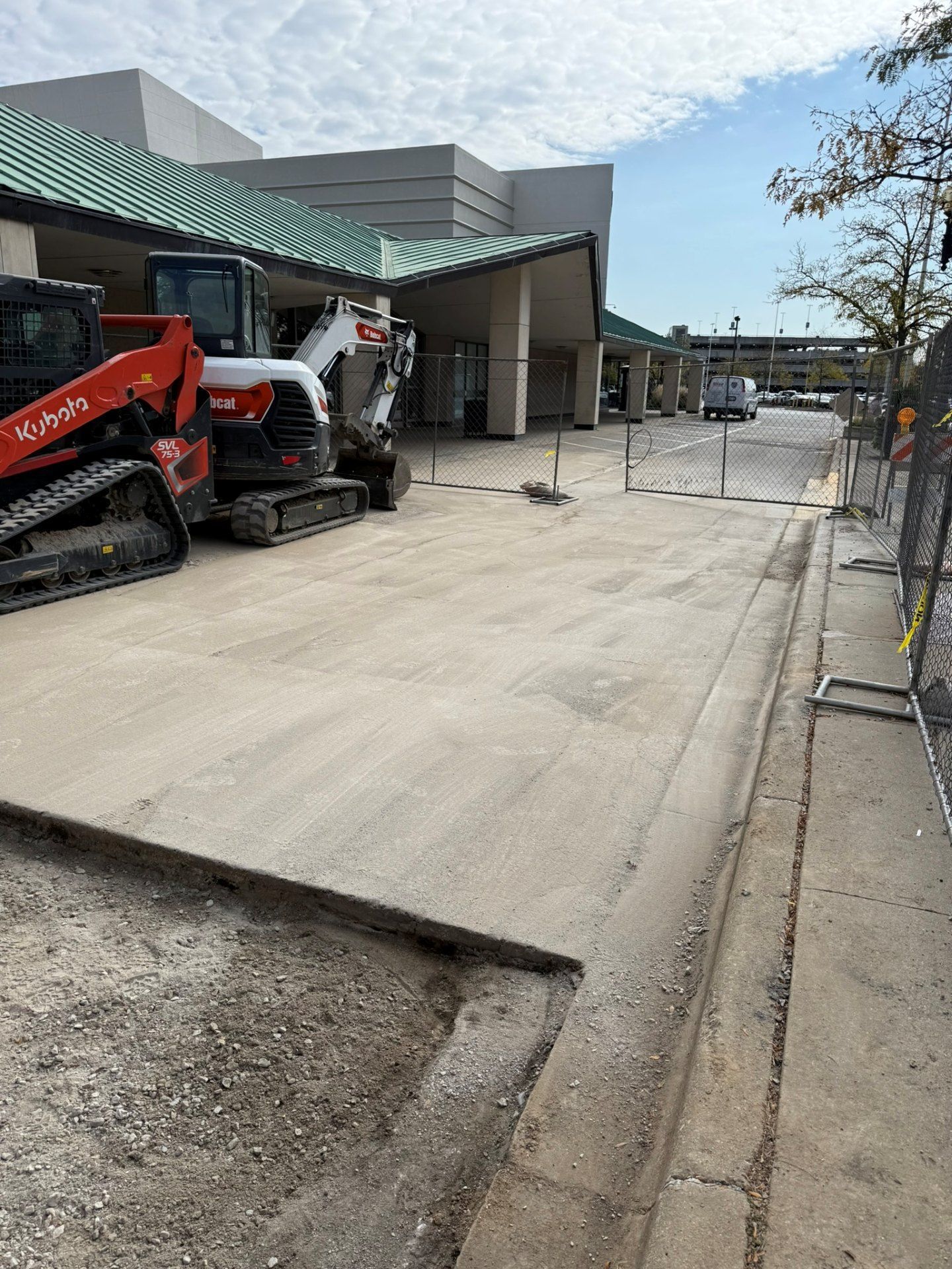 Construction site: Bobcat machinery on a concrete surface. Building with green roof in the background.