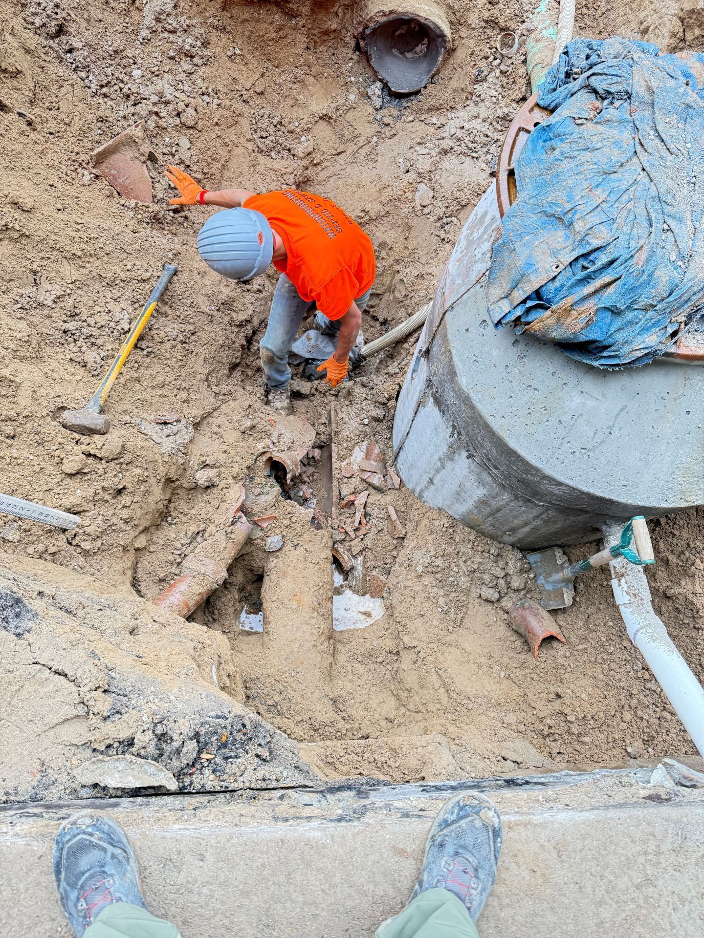 Worker in orange shirt and gloves in a dirt excavation. Concrete structure and pipes visible.