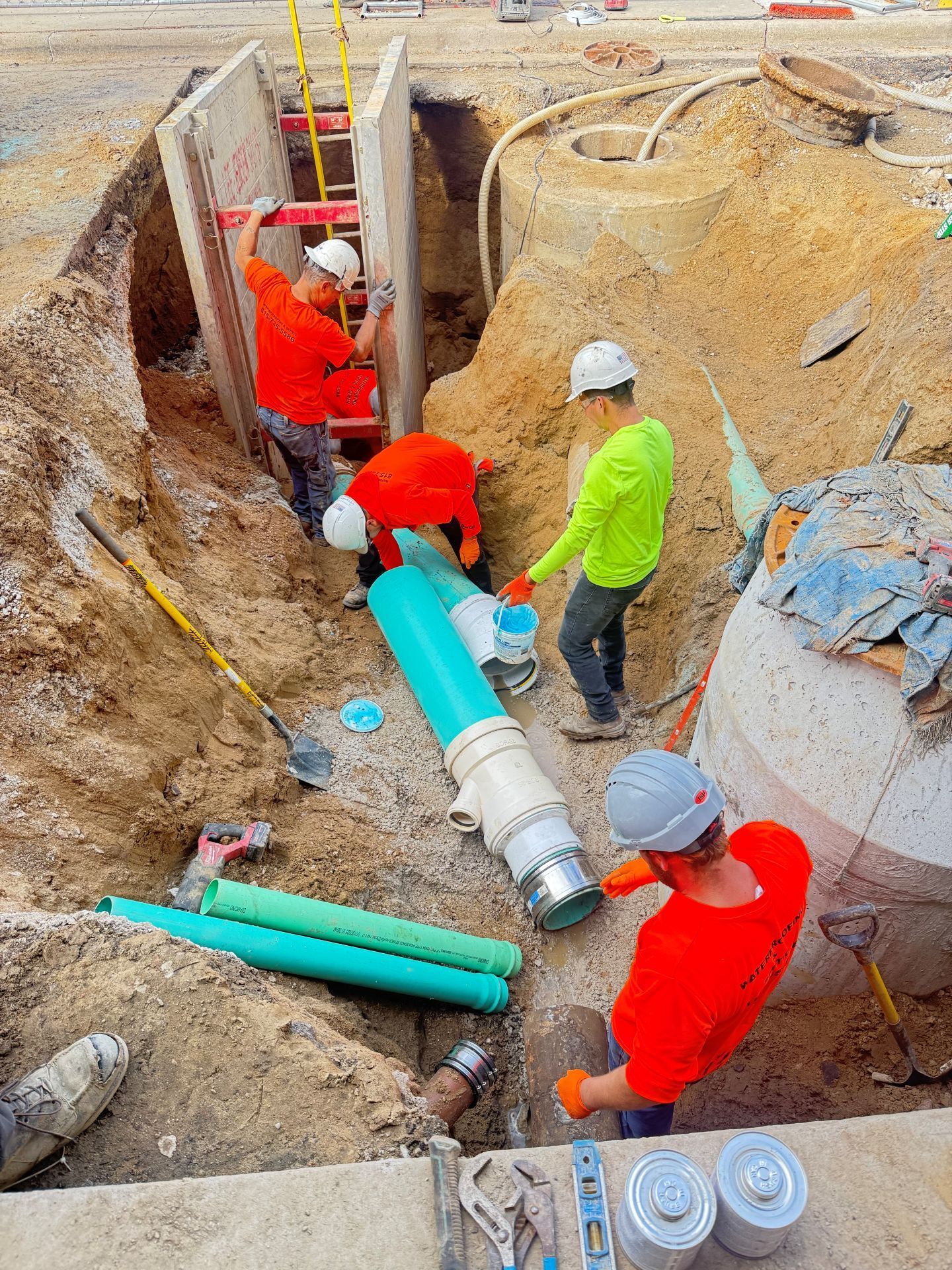 Construction workers installing pipes in a trench, using tools and wearing safety gear.