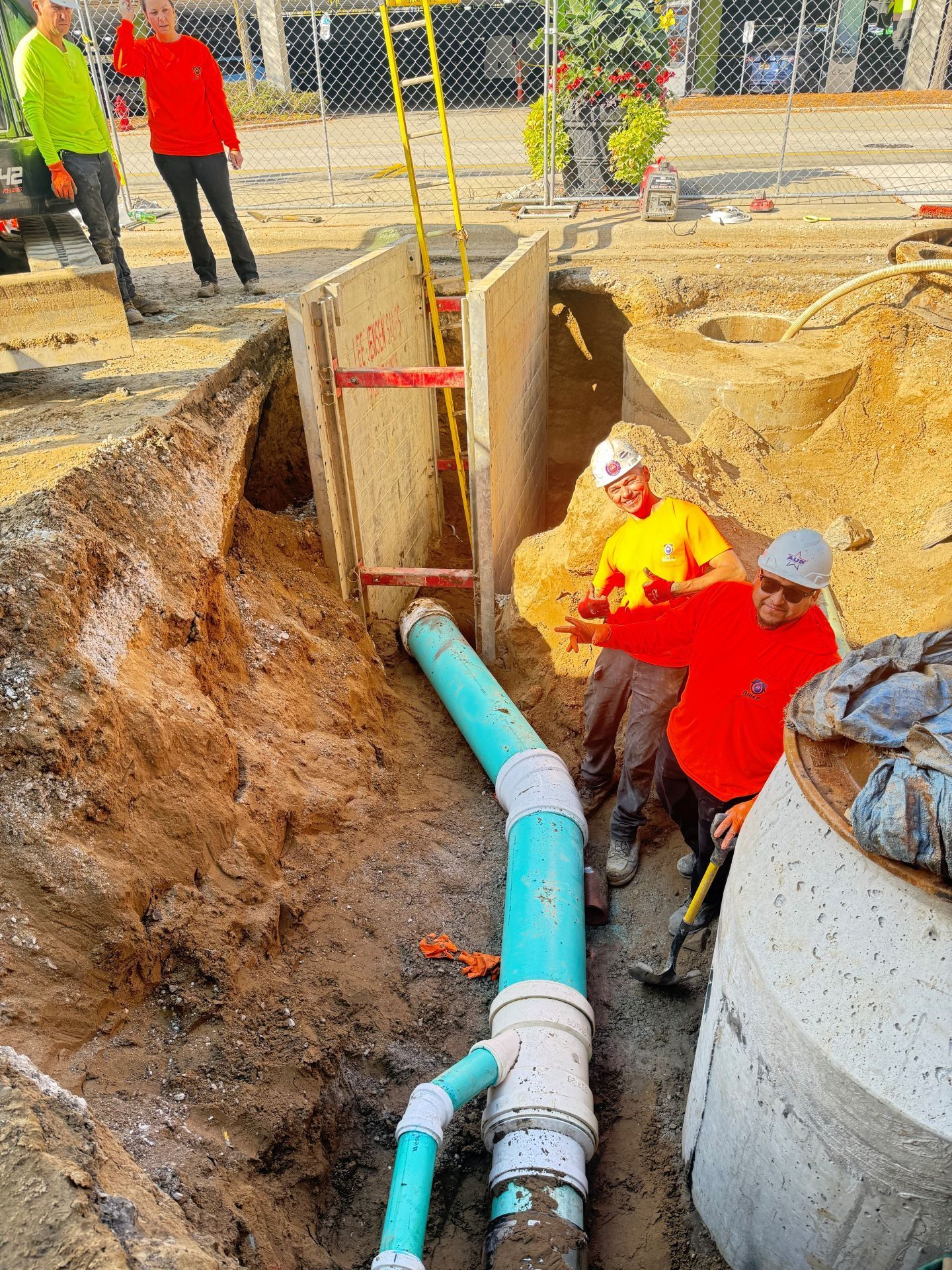 Construction workers by an open trench, installing blue pipe. Safety barriers and a ladder are present.
