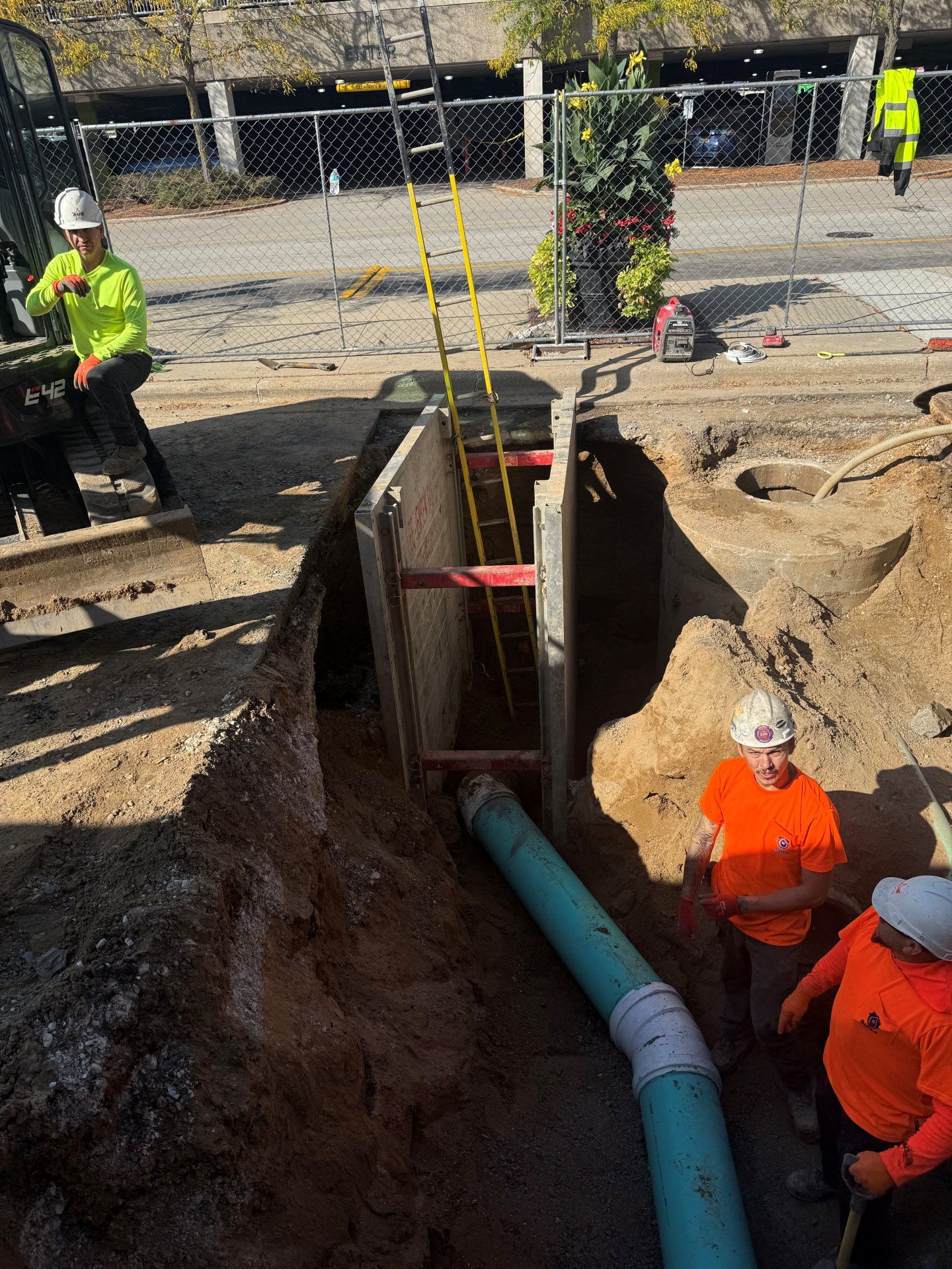 Construction workers installing a pipe in a trench. One worker operates machinery, others stand nearby.