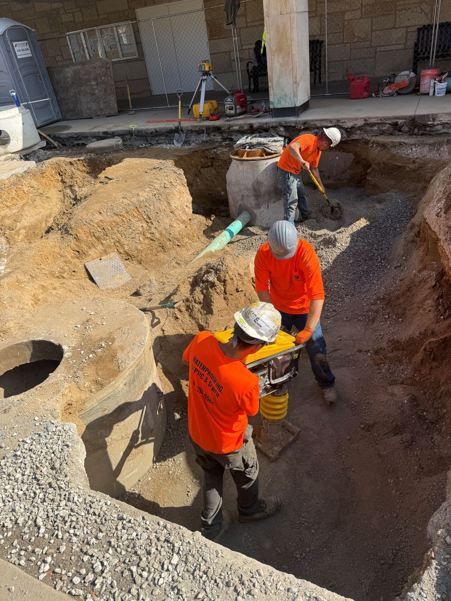 Construction workers in orange shirts excavate a site with tools near a building.