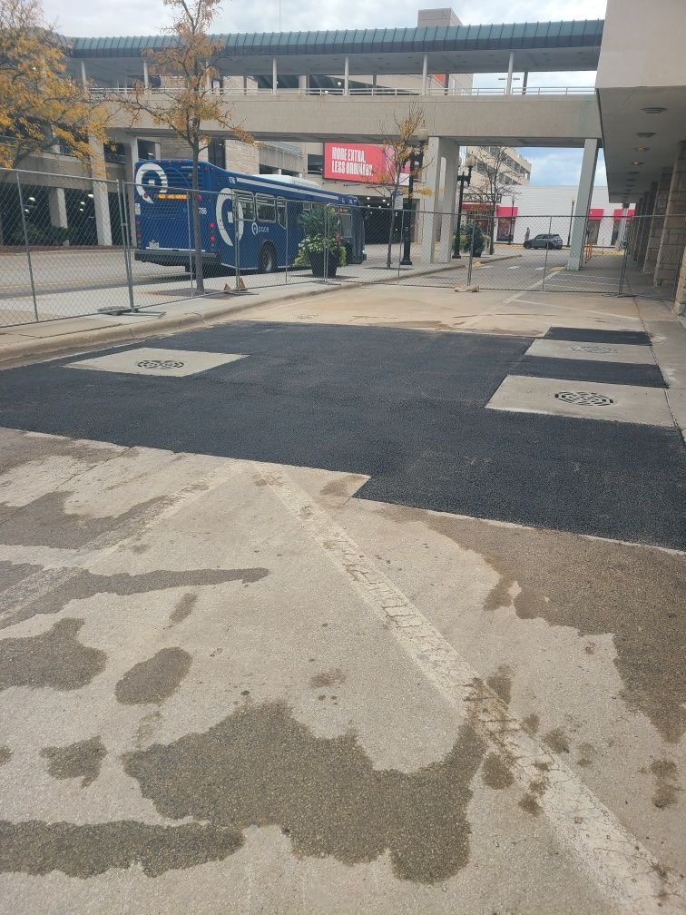 A section of freshly paved asphalt in a paved area, with a bus and a pedestrian bridge in the background.