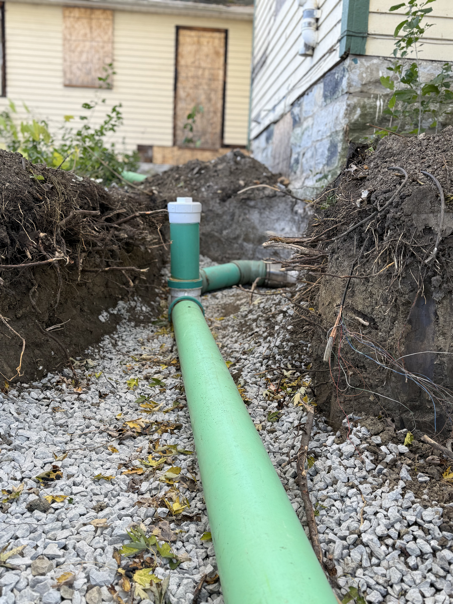 New green PVC sewer line installation in a trench beside a house foundation, with gravel base and cleanout pipe visible.