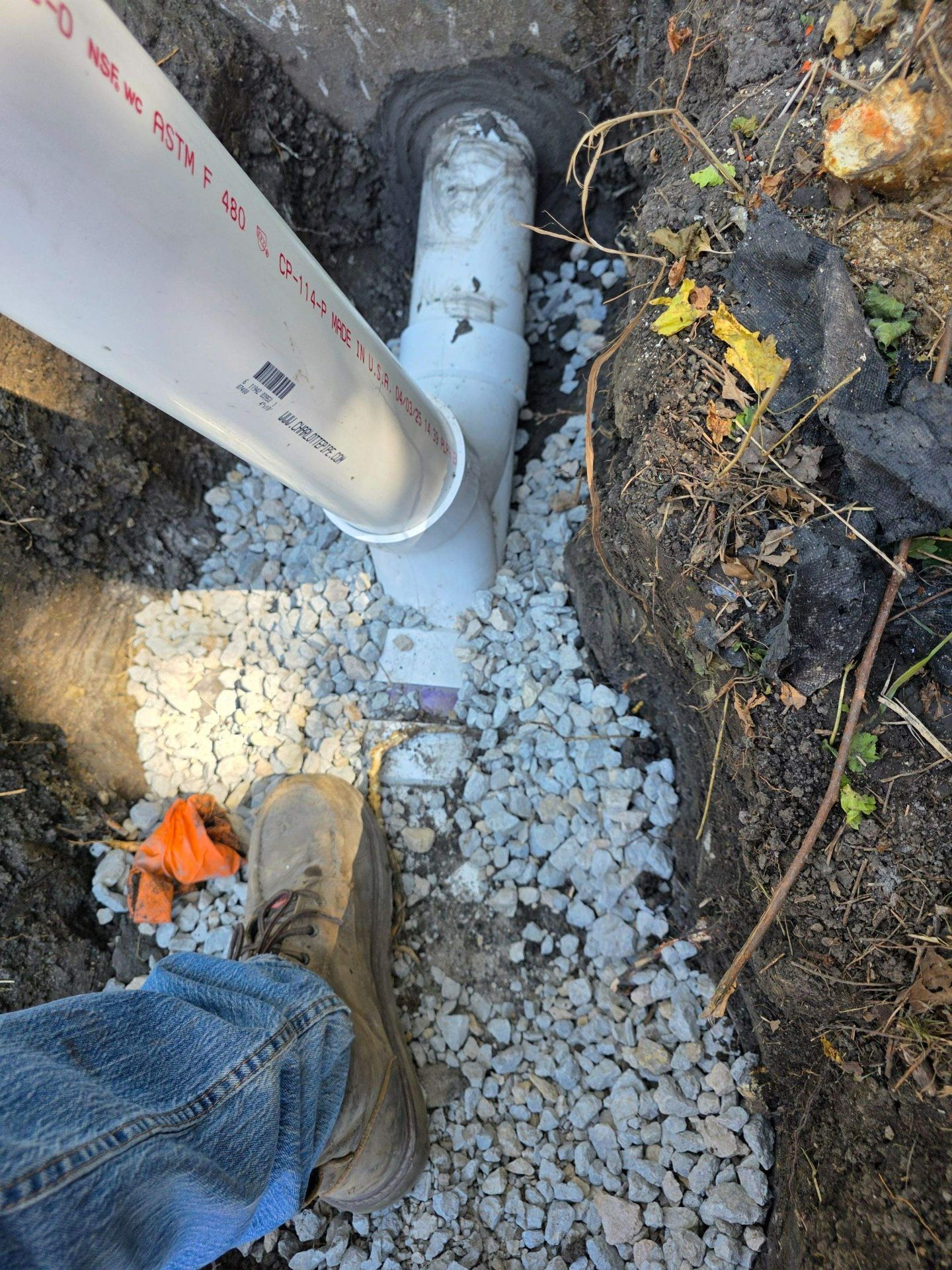 New green PVC sewer line installation in a trench beside a house foundation, with gravel base and cleanout pipe visible.