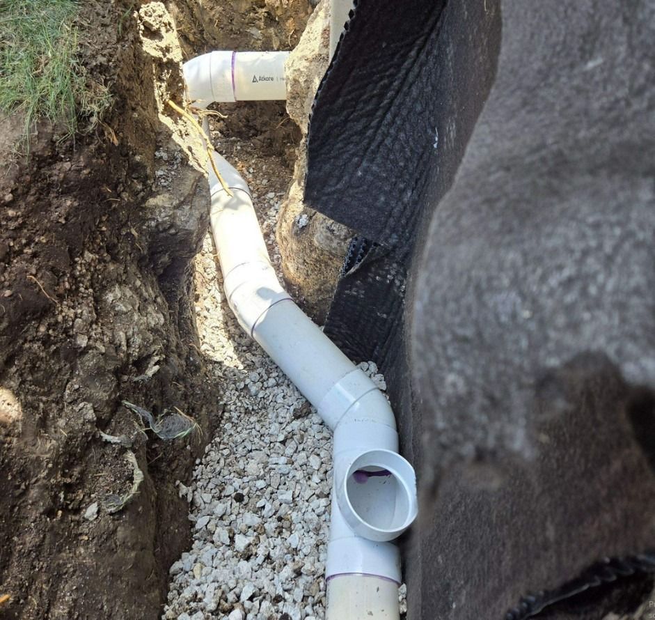White PVC drain pipes installed in a trench filled with gravel. Black fabric lines the trench.