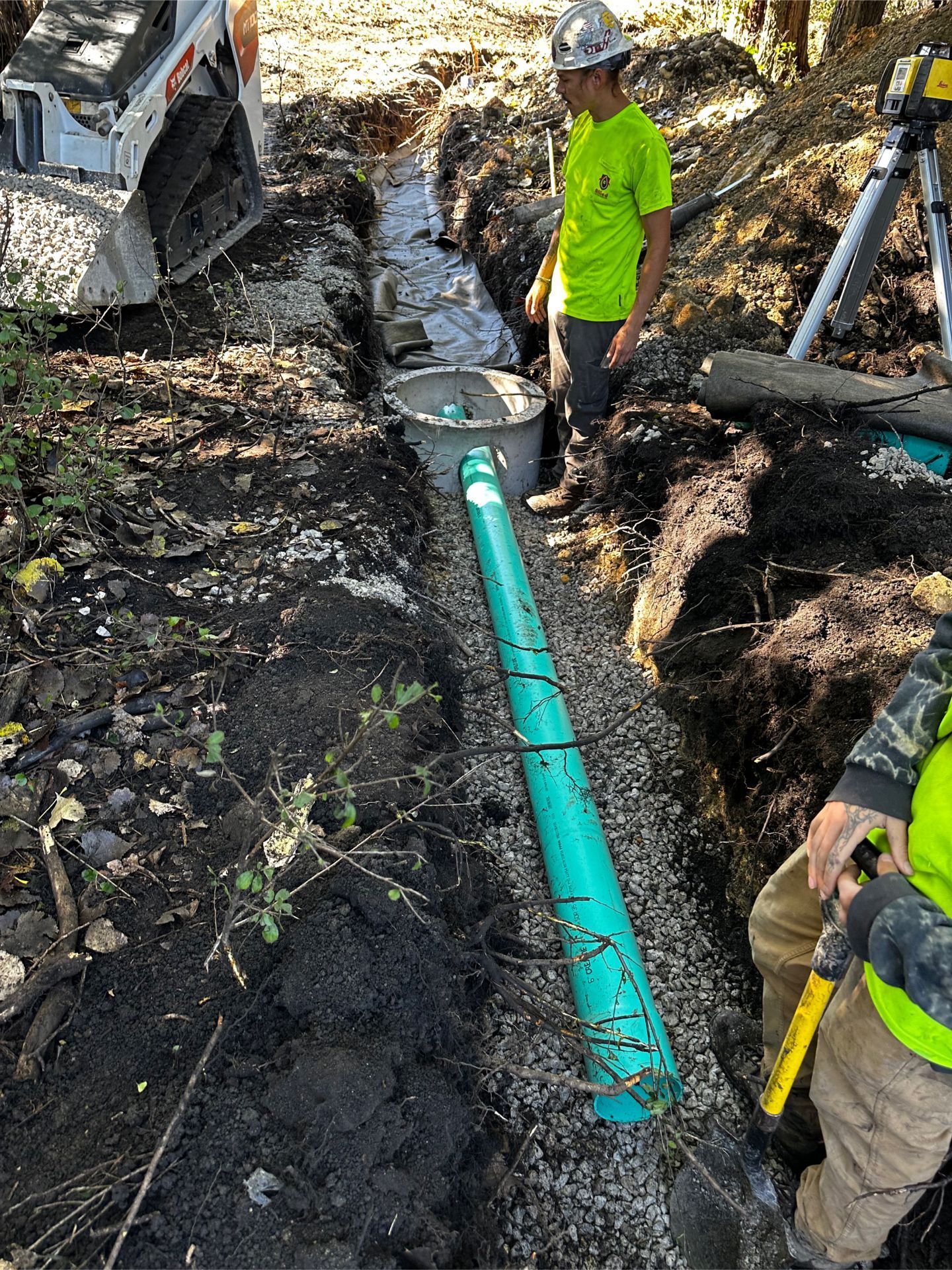 Construction workers installing a green drainage pipe in a trench filled with gravel.