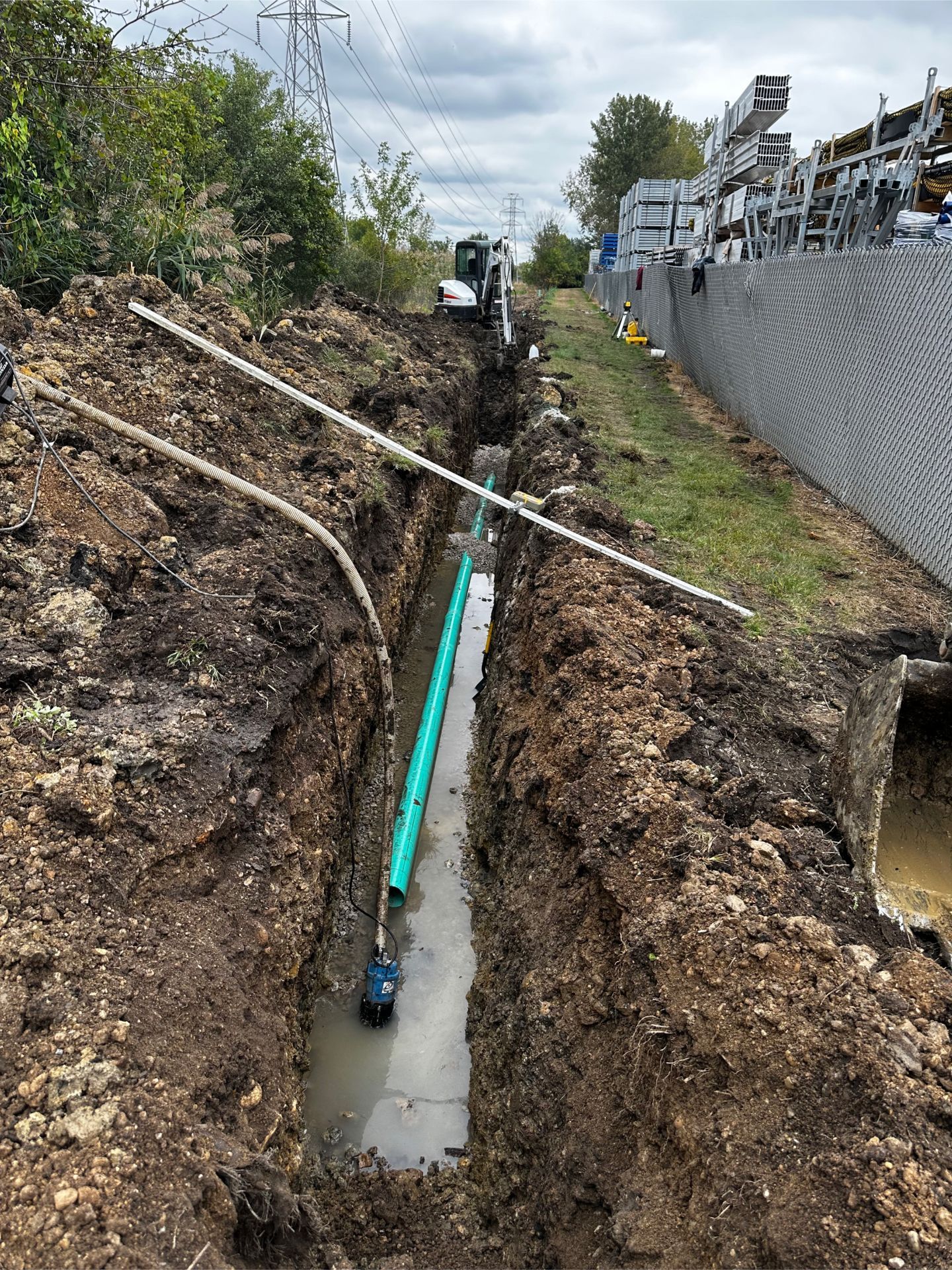 Trench dug alongside a retaining wall with a green pipe visible; machinery and utility poles in the background.