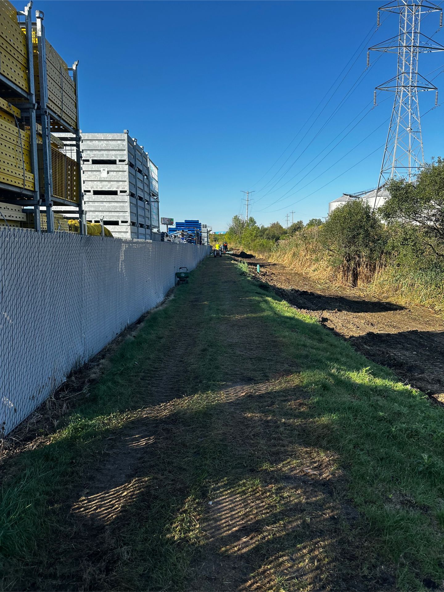 A narrow, grassy path between a fence and vegetation, with a blue sky and power lines in the background.