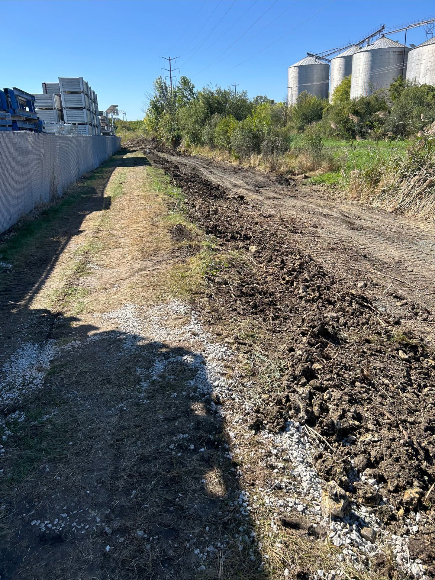 Dirt path next to a chain link fence and a plowed area, silos in the distance.