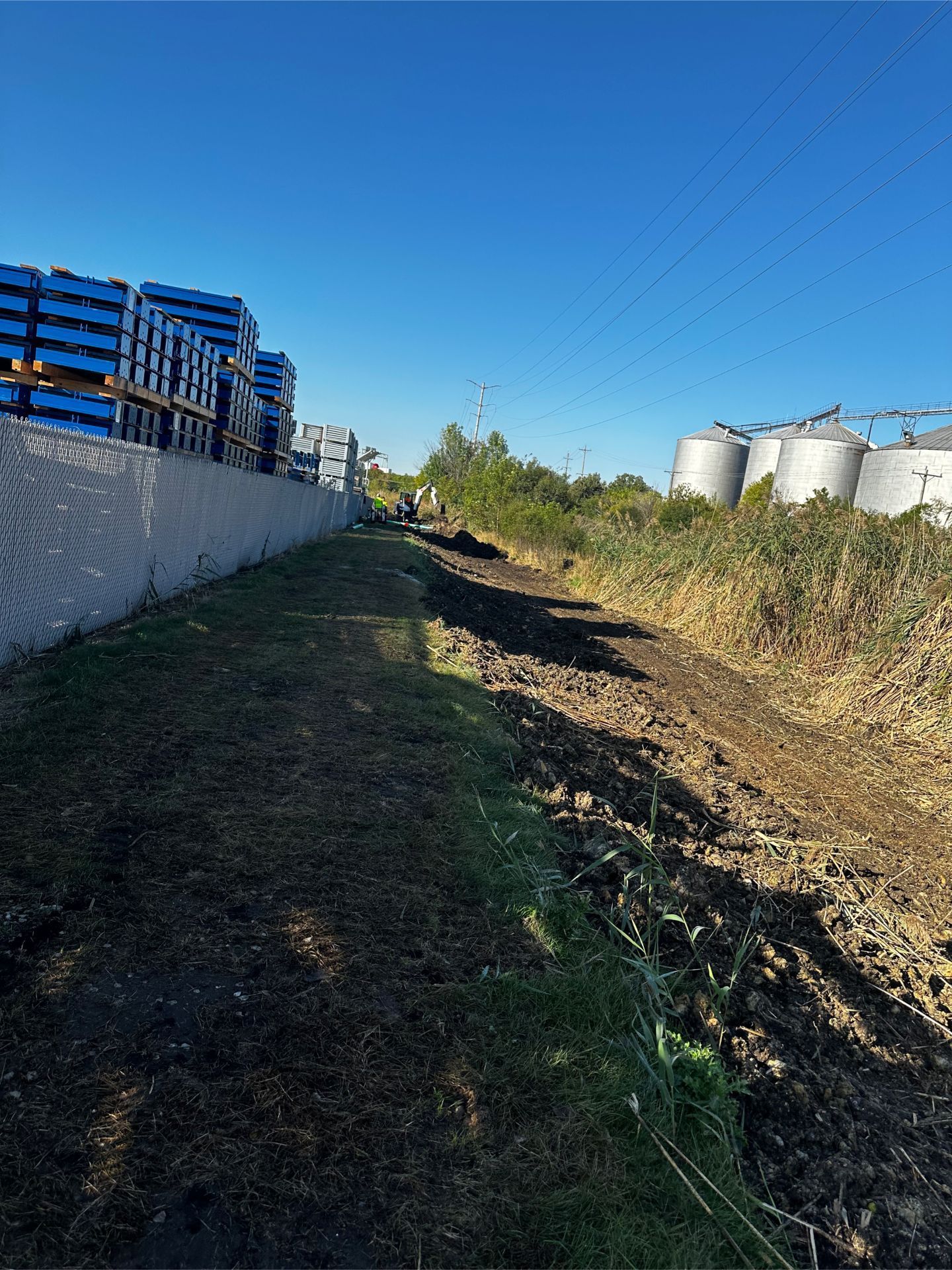 Muddy path alongside a fence and stacks of blue crates, with foliage and silos in the background under a blue sky.